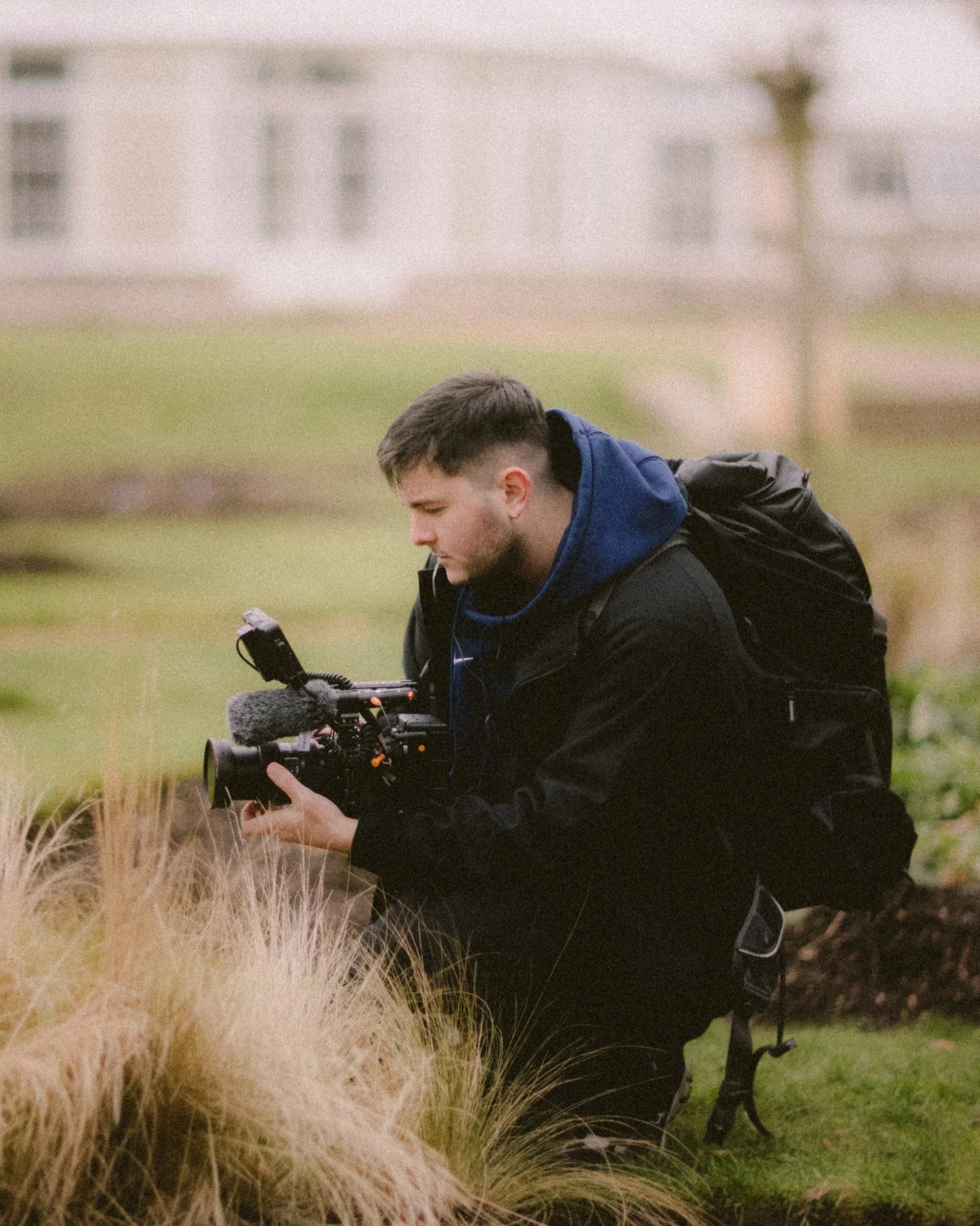 Un jeune homme avec un sac à dos noir, vêtue d'une veste noire et d'une chemise bleue, tient une caméra professionnelle tout en étant accroupi dans un espace extérieur avec de l'herbe et des plantes décoratives, devant un bâtiment en arrière-plan.