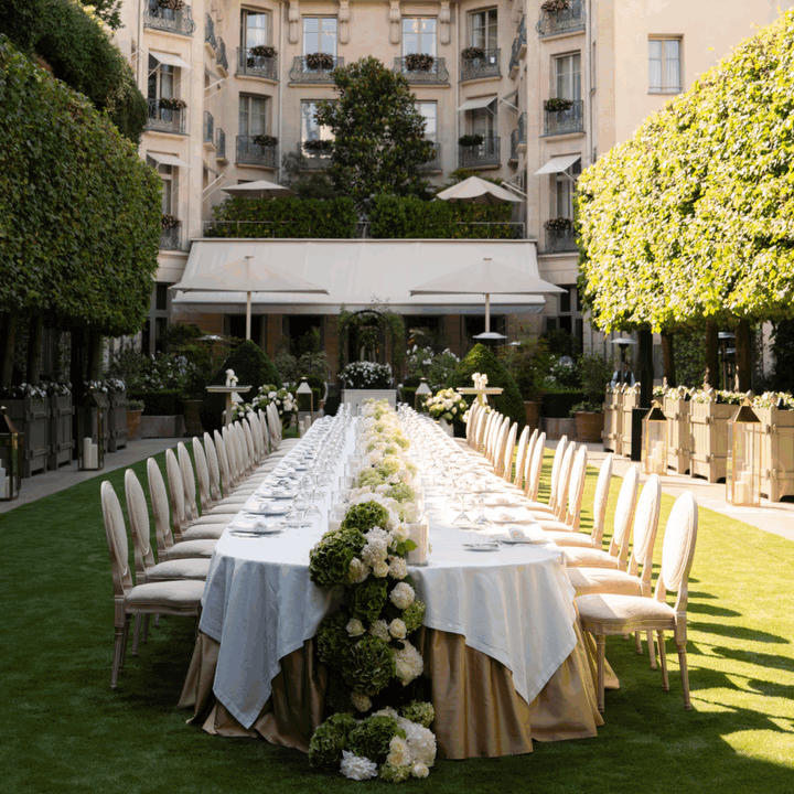 Table dressée pour un événement dans un jardin, surrounding des chaises et décorations florales, avec un bâtiment en arrière-plan.