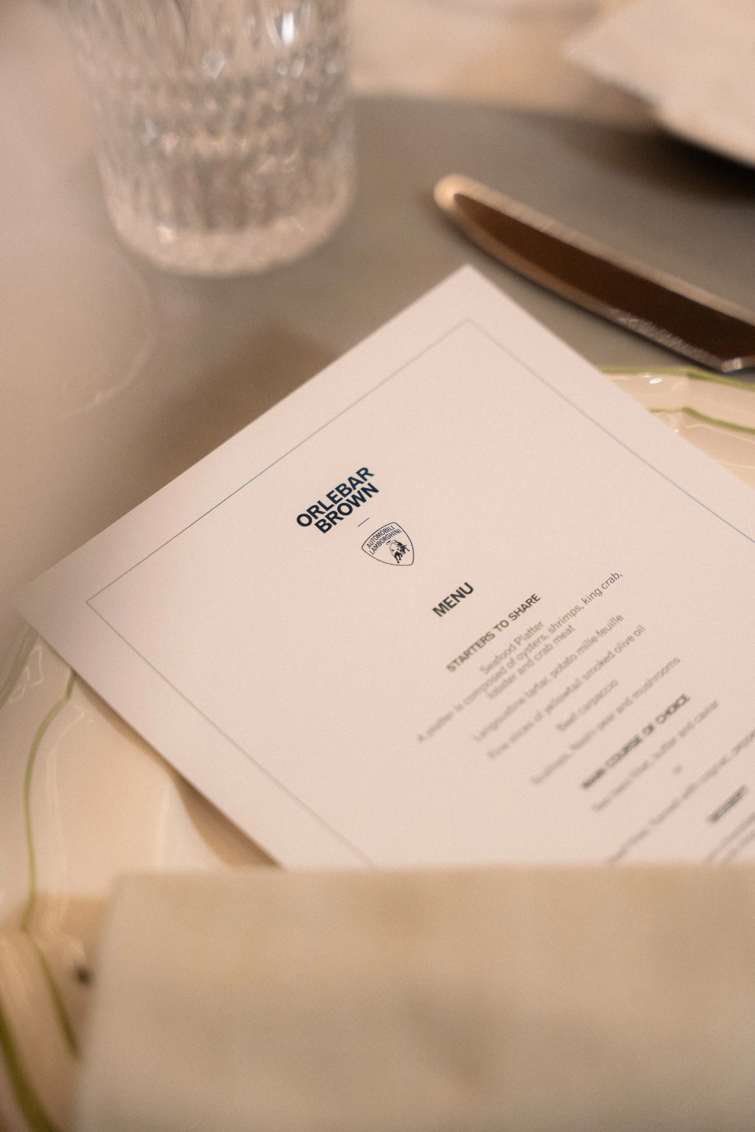 Menu placé sur une table, avec un verre en verre taillé, un couteau et des assiettes. Le menu est de l'hôtel Orlebar Brown avec un logo Lamborghini au sommet.
