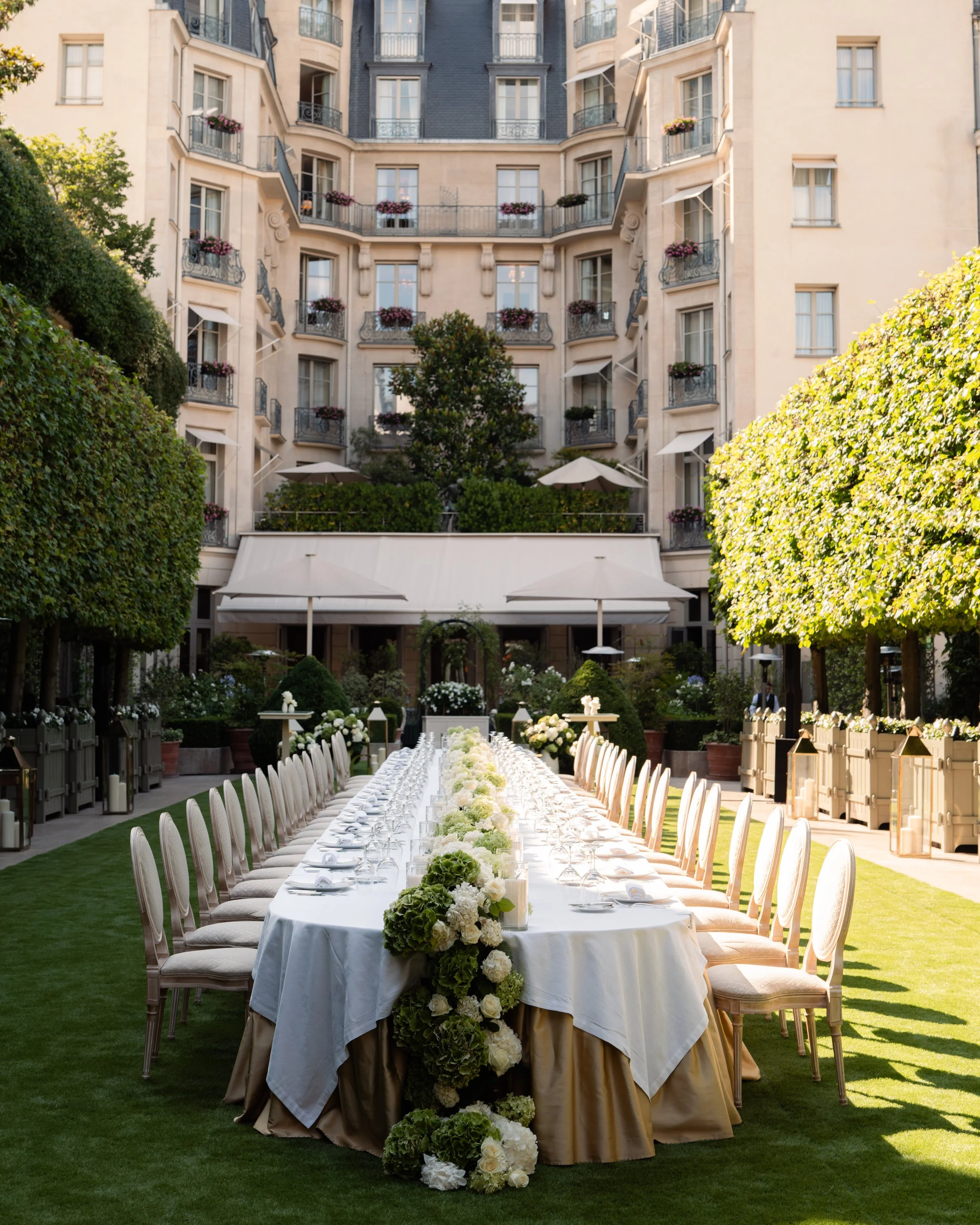 Table de banquet dressée dans un jardin en plein air, entourée d'arbres et de bâtiments résidentiels, avec fleurs et décorations pour un événement.
