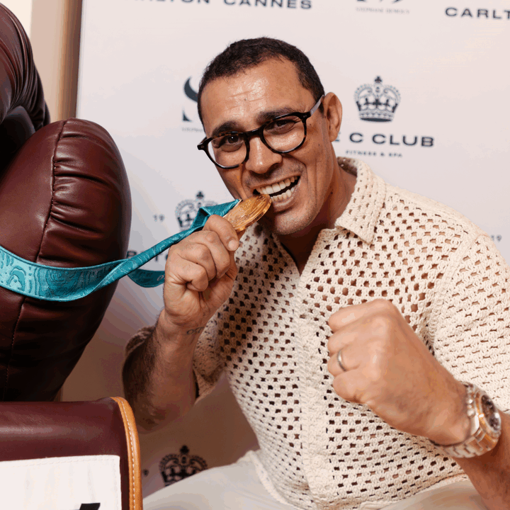 Un homme souriant mordant une médaille lors d'une cérémonie ou d'une réception, avec un fond comportant des logos.