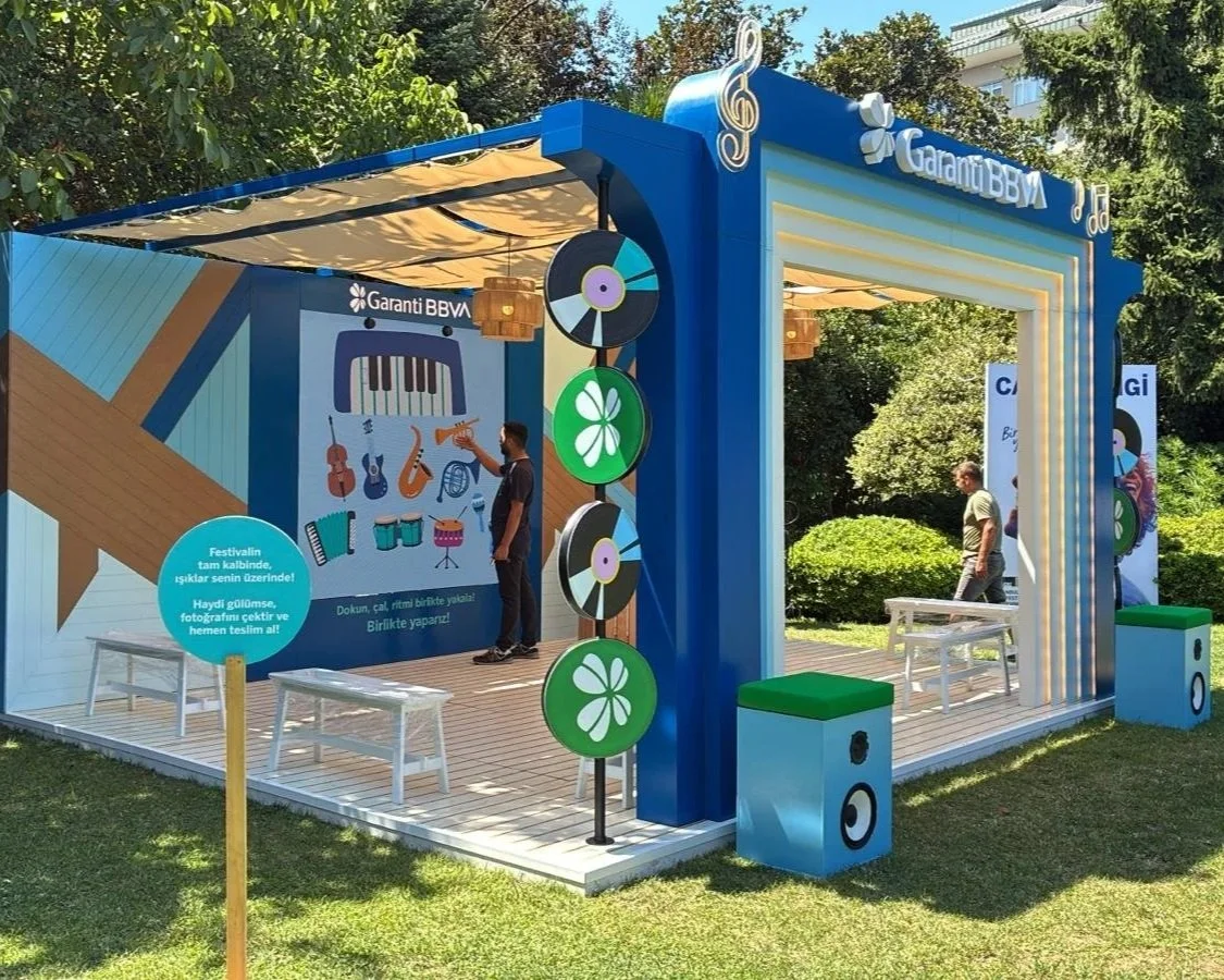 Müzik temalı açık hava etkinliği alanı, içinde enstrüman resimleri ve paneller ile süslenmiş, iki kişi etrafında birkaç kişi ve ağaçlar var.