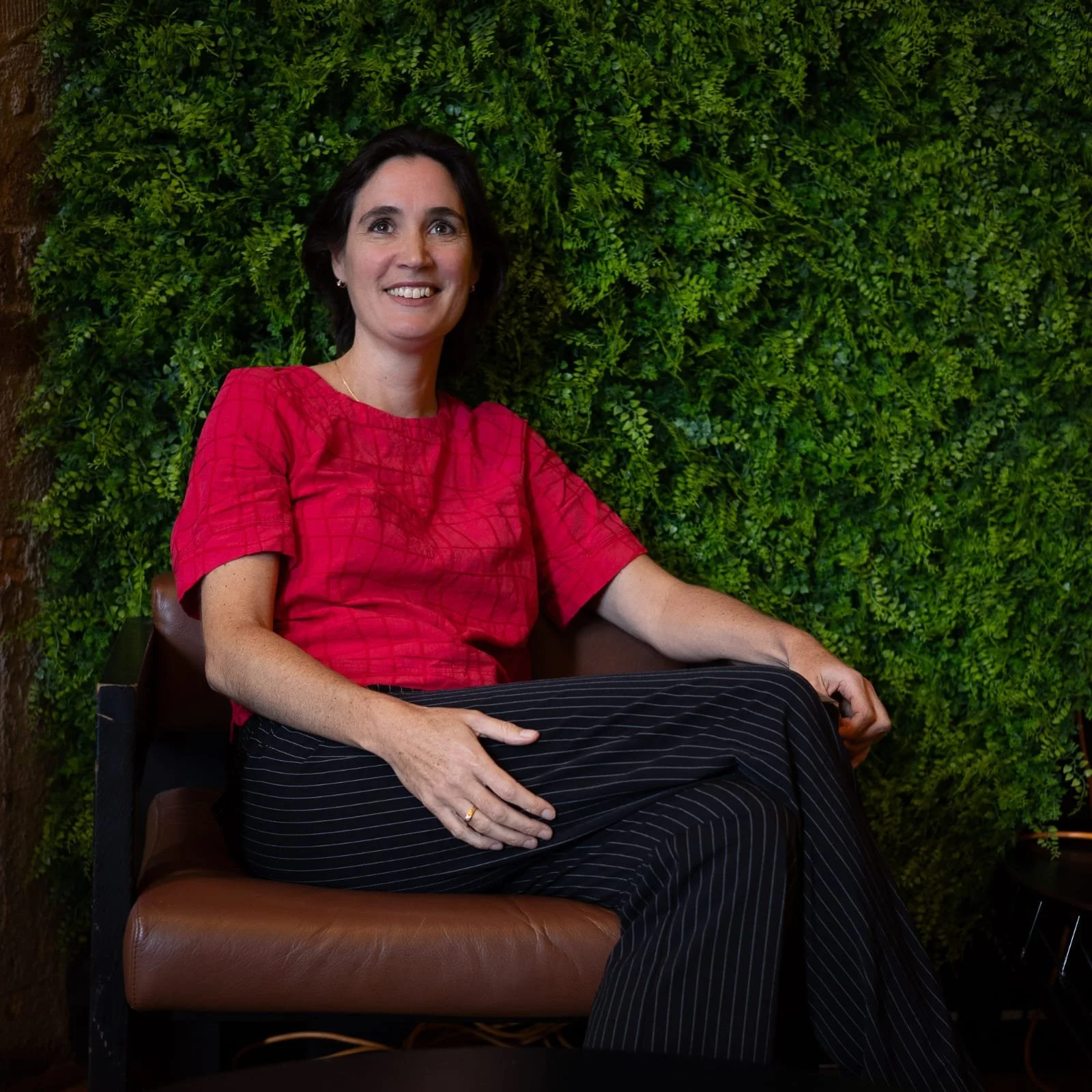 A woman sitting on a brown chair in front of a green leafy wall, smiling, wearing a red blouse and black pinstripe pants.