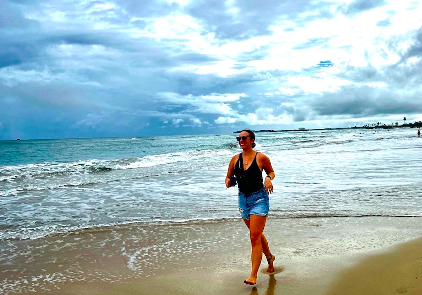 A woman in sunglasses, a black tank top, and denim shorts walking along the beach with the ocean and cloudy sky in the background.