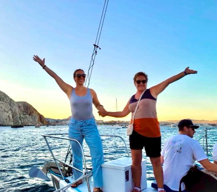 Two women are standing on a boat holding hands, with arms raised and smiling. They are in a marina during sunset, with other boats and rocky cliffs in the background.
