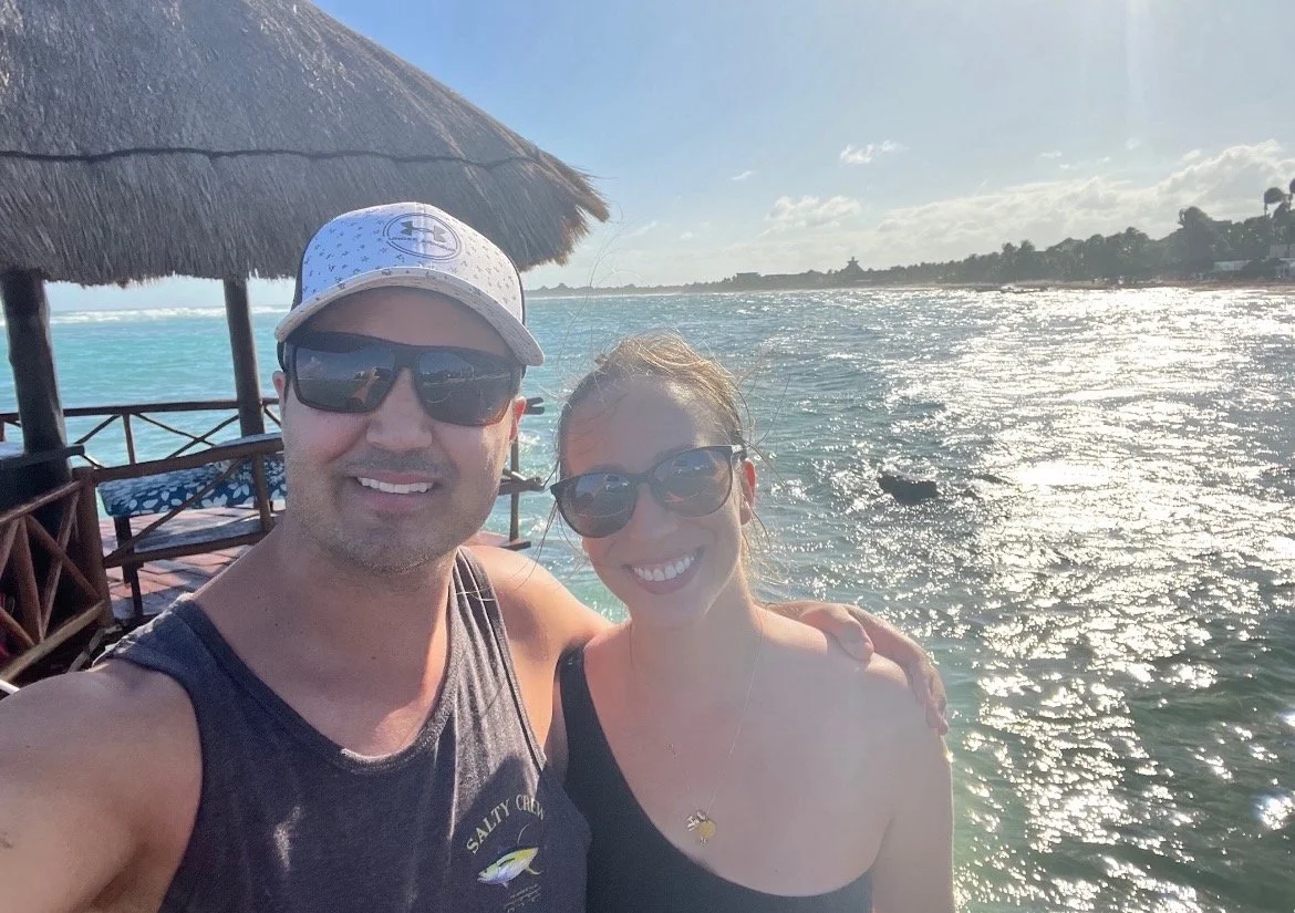 A man and woman wearing sunglasses taking a selfie by the water on a sunny day, with a thatched roof structure behind them.