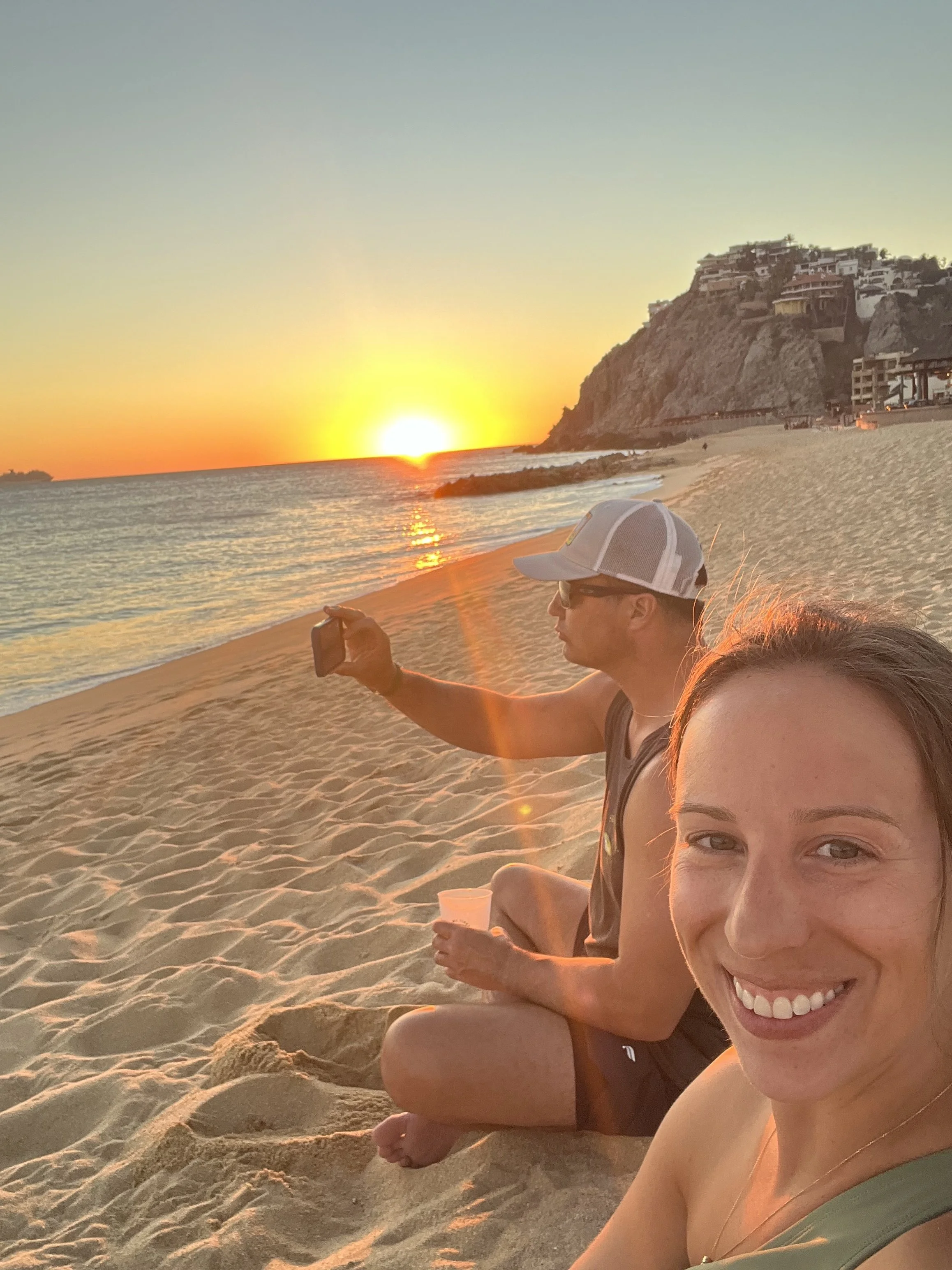 Smiling woman taking a selfie at the beach during sunset, with a man sitting beside her holding a phone, and a hillside with white buildings in the background.