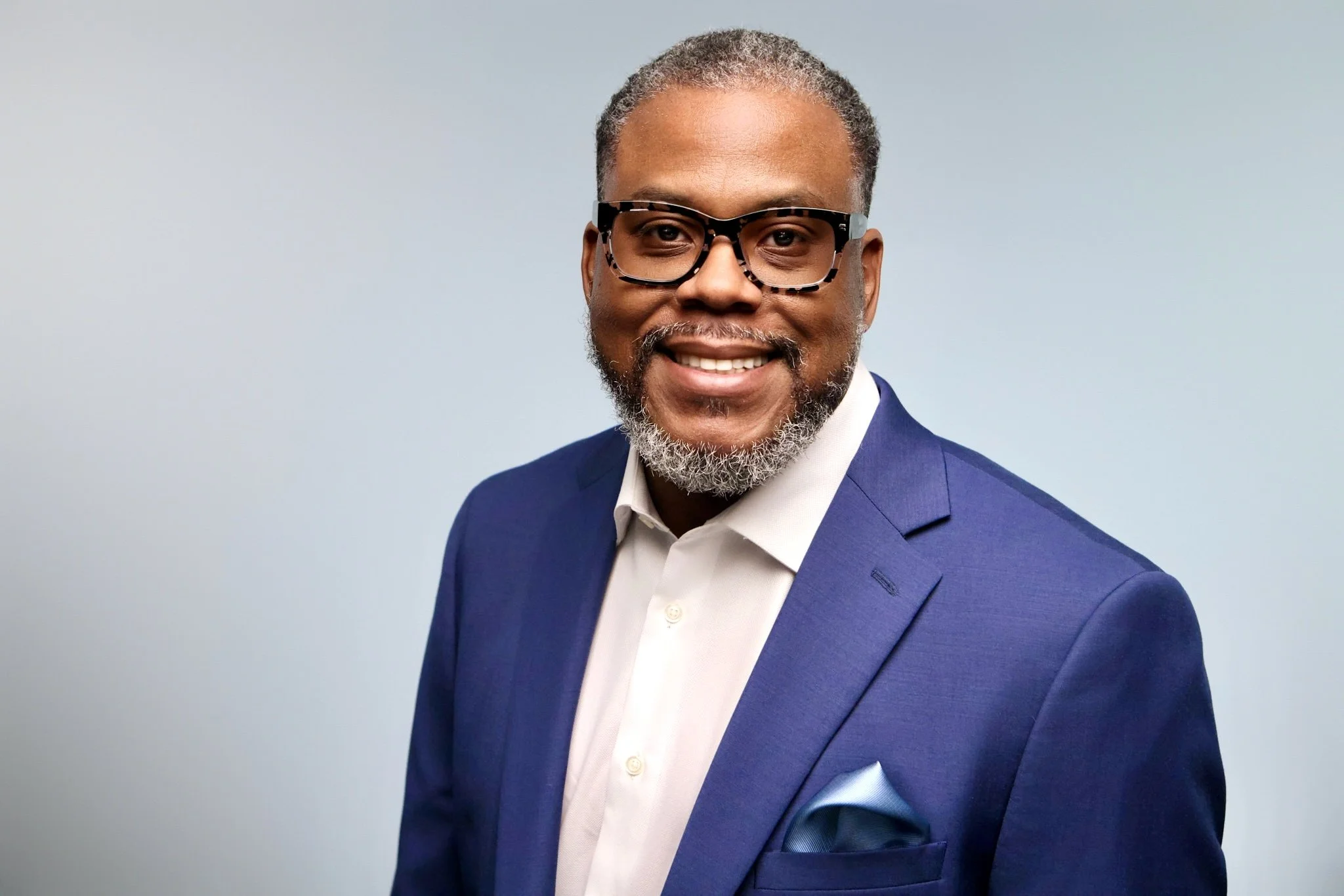 Portrait of a Black man with gray hair and beard, wearing glasses, a white shirt, and a blue suit jacket, smiling against a light blue background.