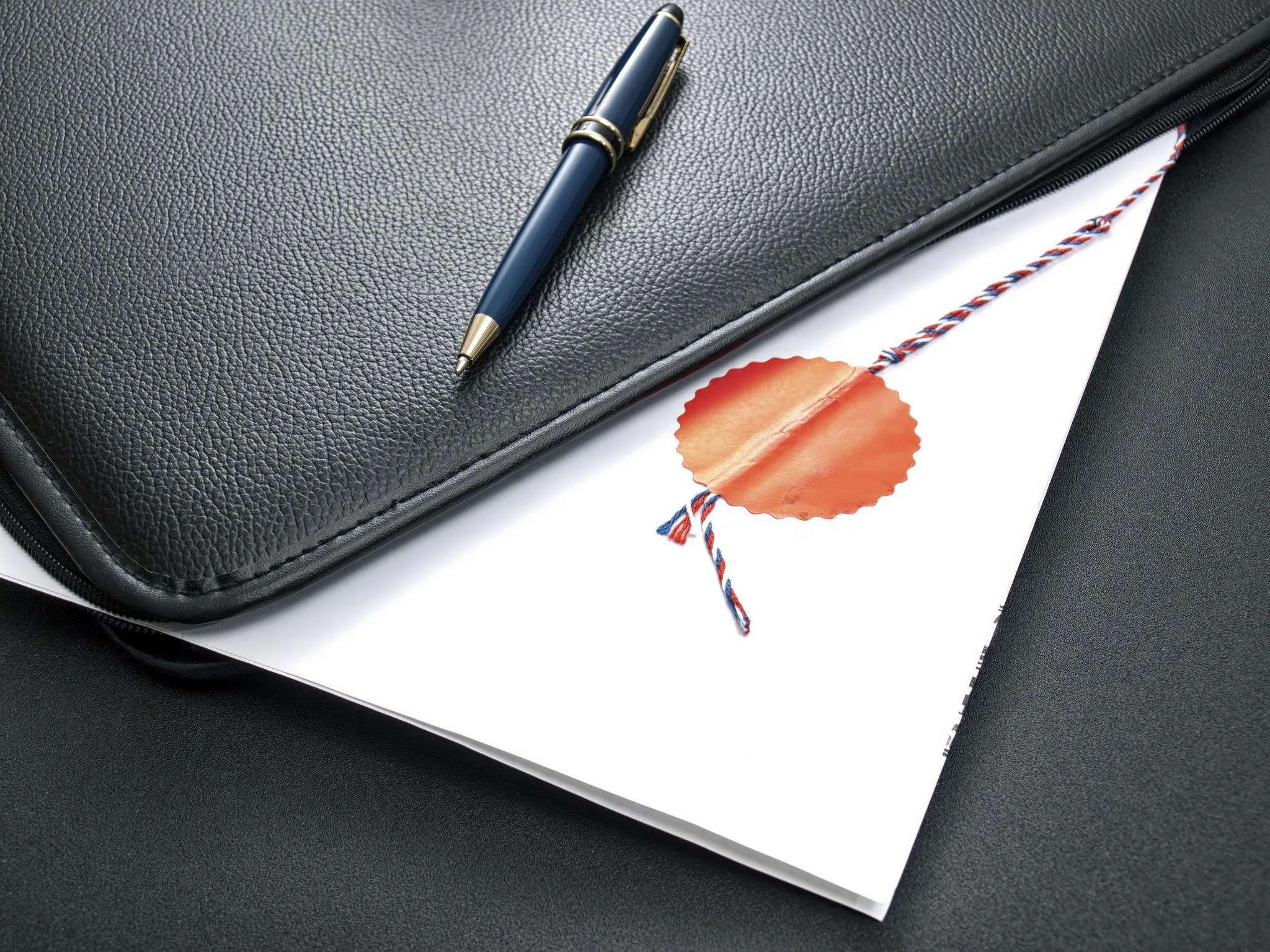 Close-up of a black textured leather briefcase with a closed zipper, a blue and gold pen resting on top, a white sheet of paper, a red and blue cord, and an orange wax seal.
