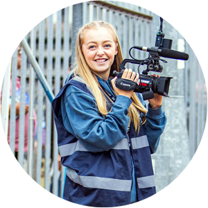 A young woman outdoors holding a professional video camera, smiling, wearing a blue raincoat.