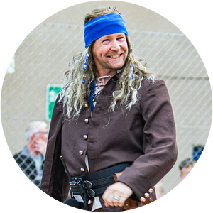 Man with long hair and a beard wearing a blue bandana and brown coat, smiling outdoors.