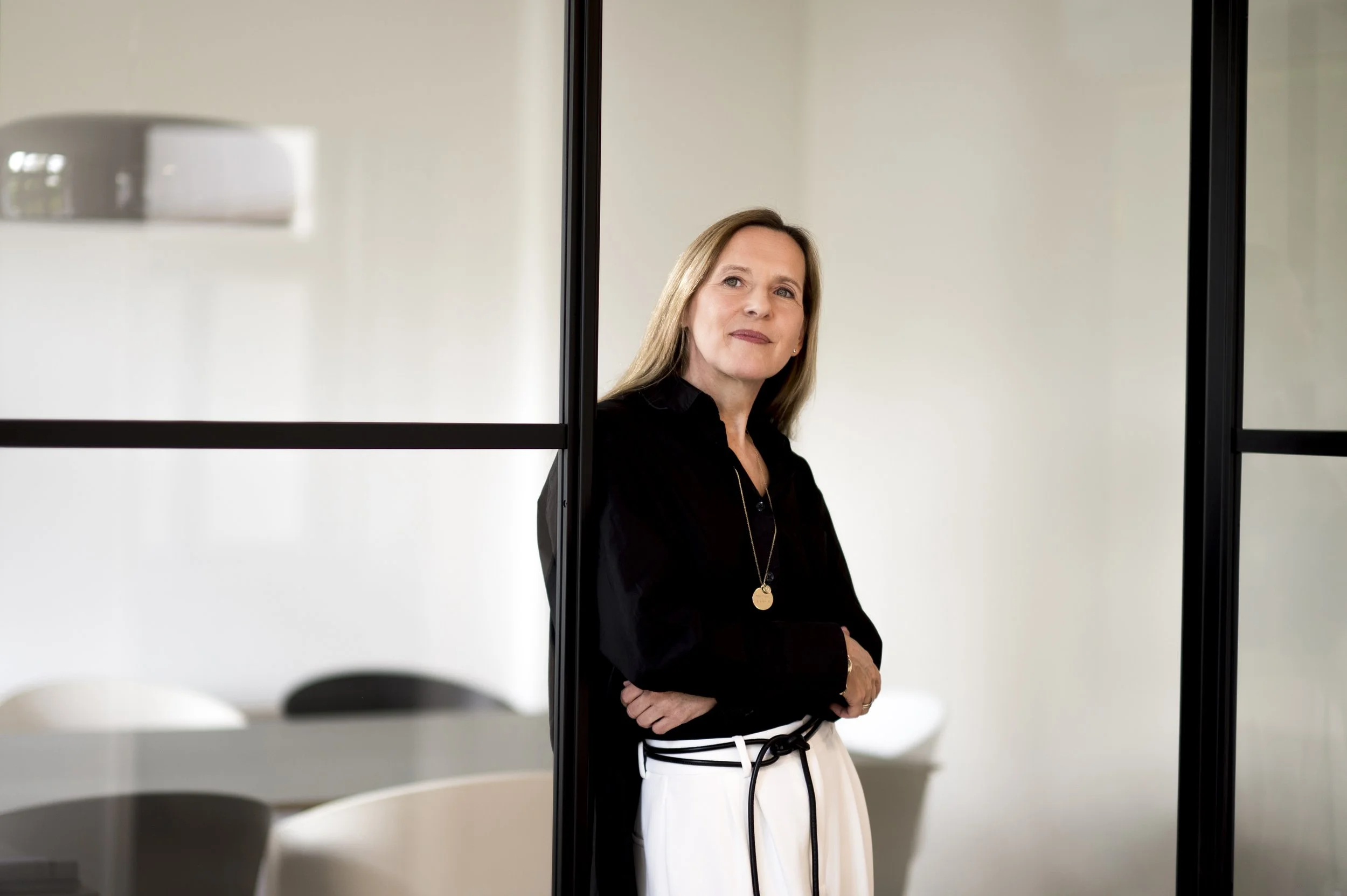 Frau in einem modernen Büro läuft an einer Glaswand vorbei, trägt schwarze Bluse und weiße Hose, wirkt nachdenklich.