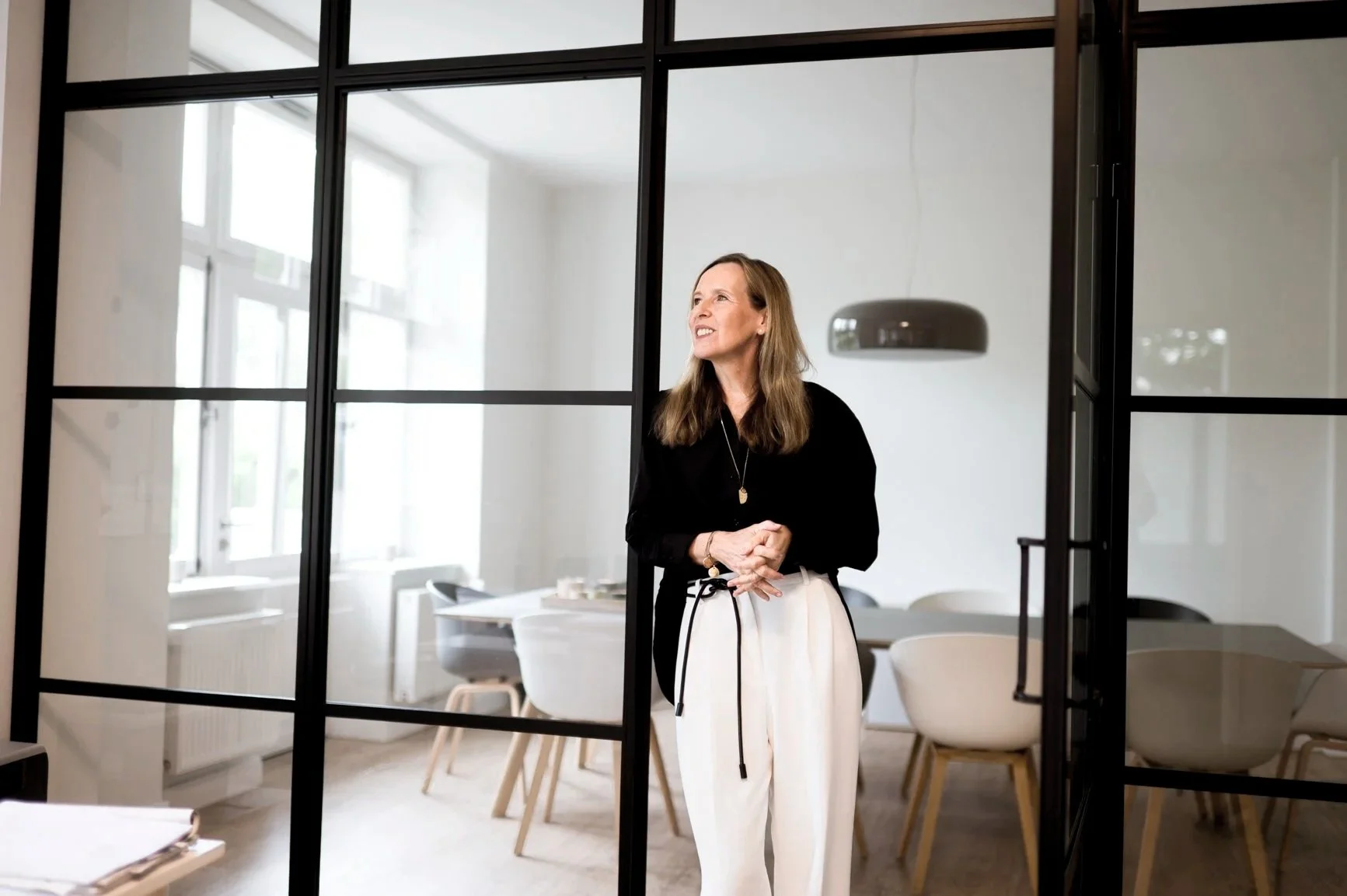 Eine Frau steht in einem modernen Konferenzraum, sichtbar durch eine Glaswand, lächelt und schaut zur Seite.