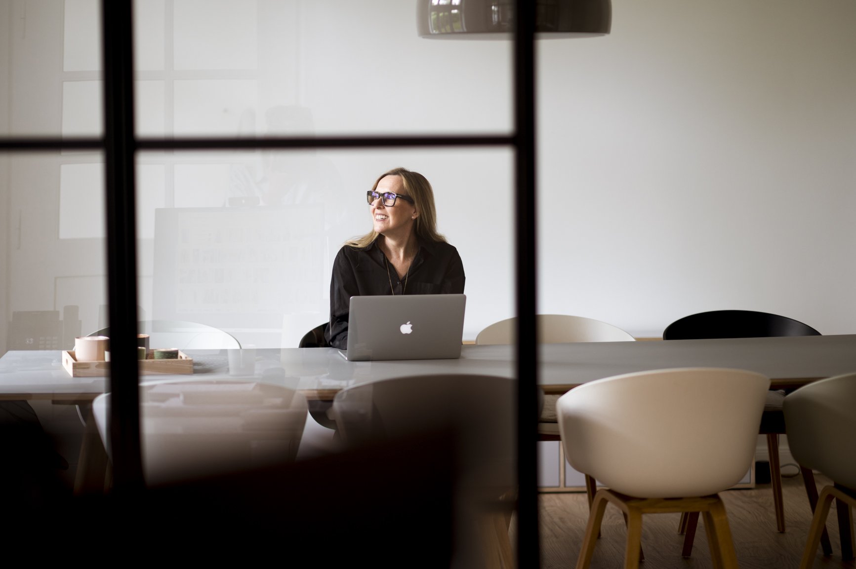 Eine Frau sitzt an einem Esstisch mit einem offenen Laptop, lächelnd und blickt zur Seite, hinter einer halbtransparenten Scheibe in einem modernen Büro.