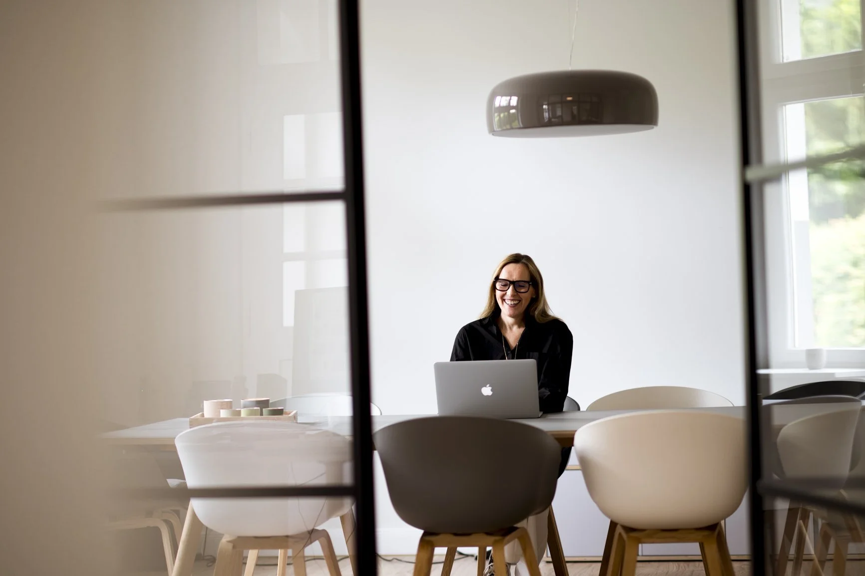 Eine lachende Frau mit Brille sitzt an einem Tisch mit einem Laptop, im Hintergrund ein weißer Raum mit großen Fenstern, durch die grünes Laub sichtbar ist.