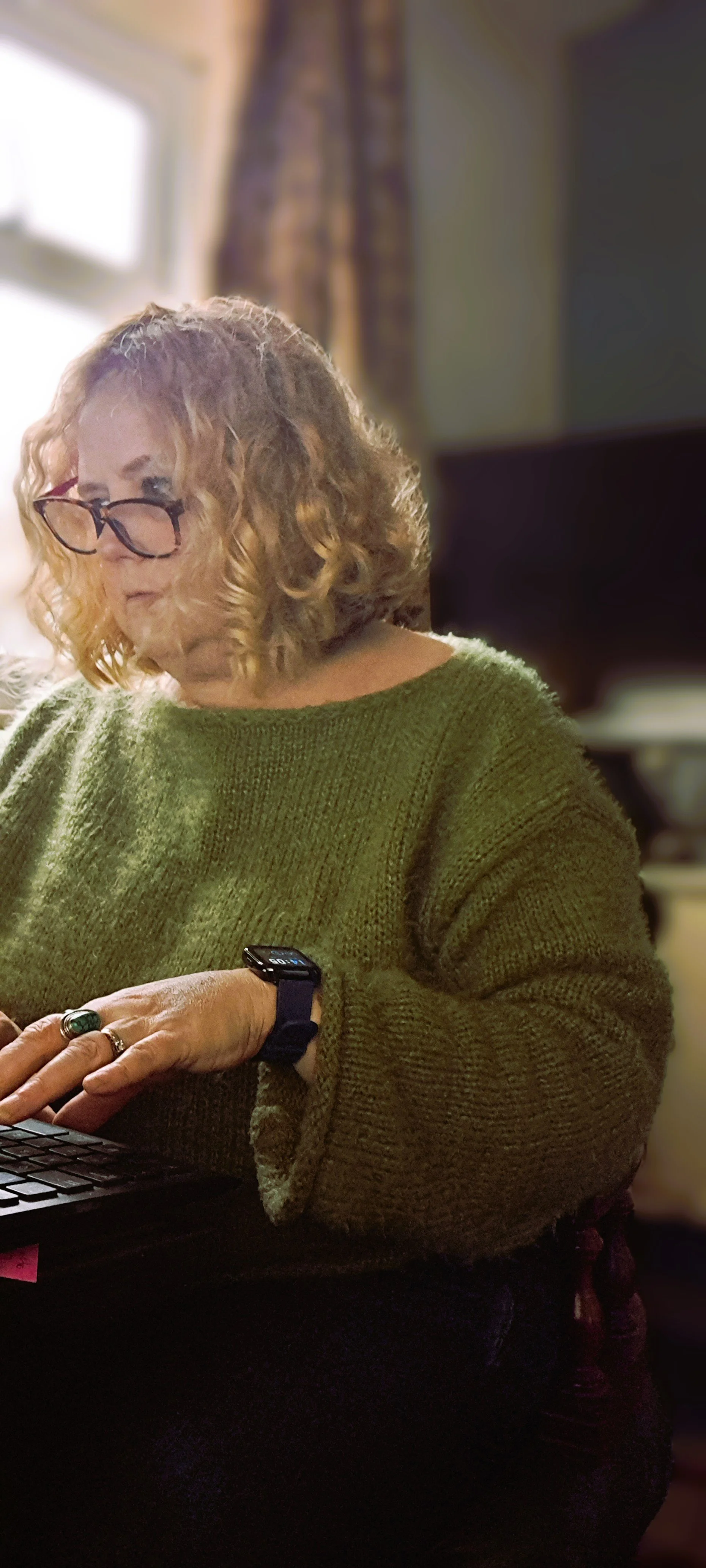Historian Emma Kay using a smartwatch and typing on a laptop in a room with a window and curtain.
