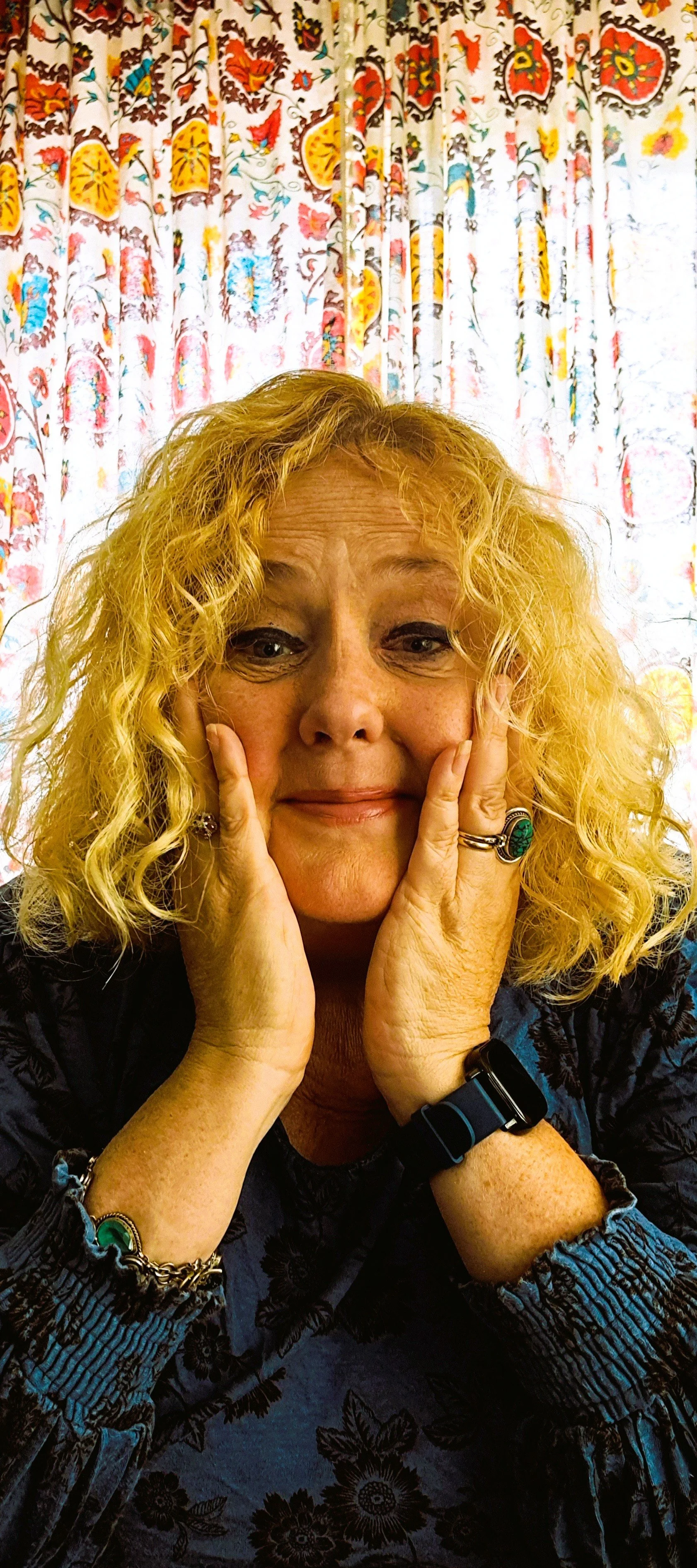 A woman with blonde, curly hair rests her face in her hands, smiling gently, seated in front of colorful, patterned curtains.