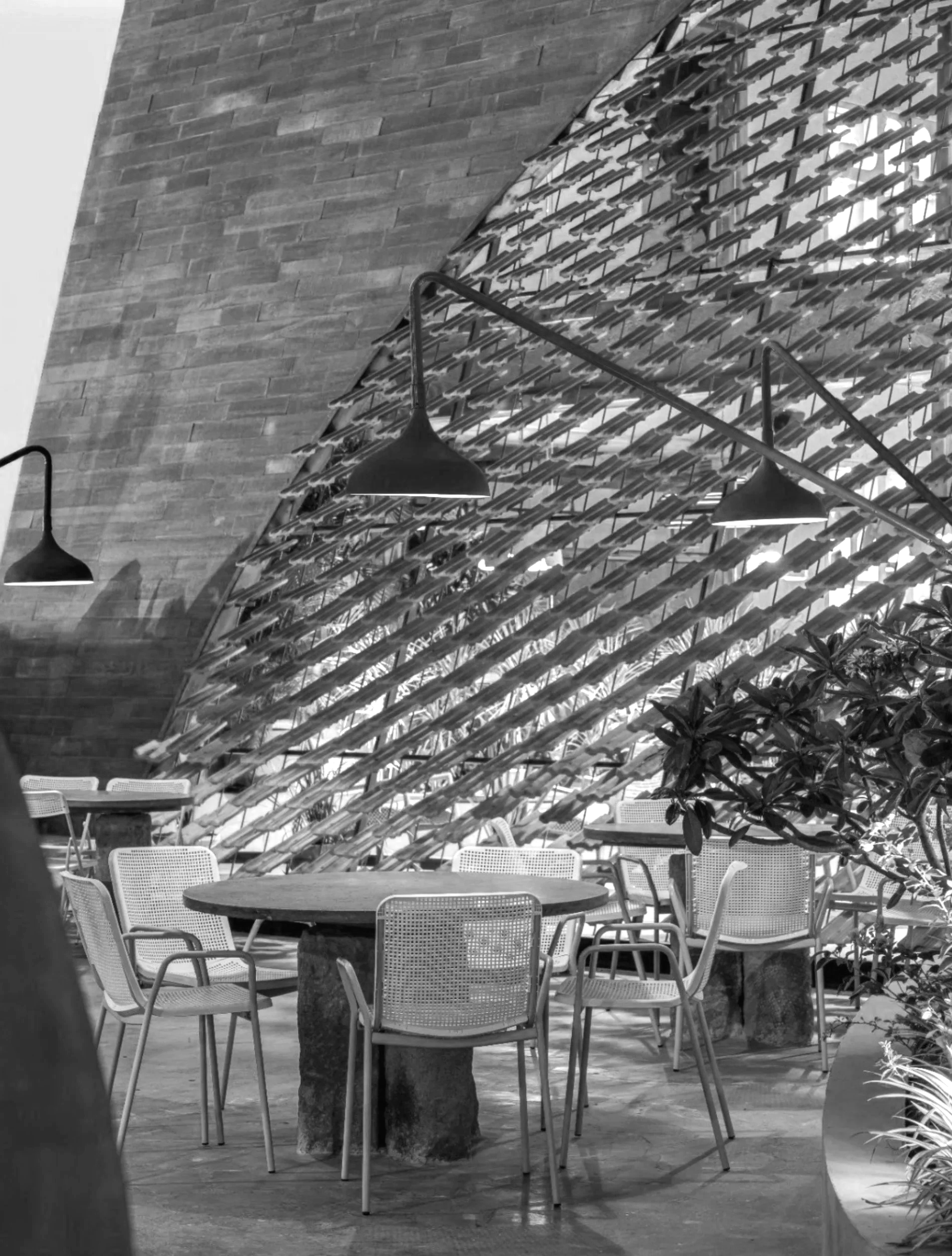 Interior of a modern building with a slanted glass ceiling, hanging light fixtures, a round table with chairs, and some plants.