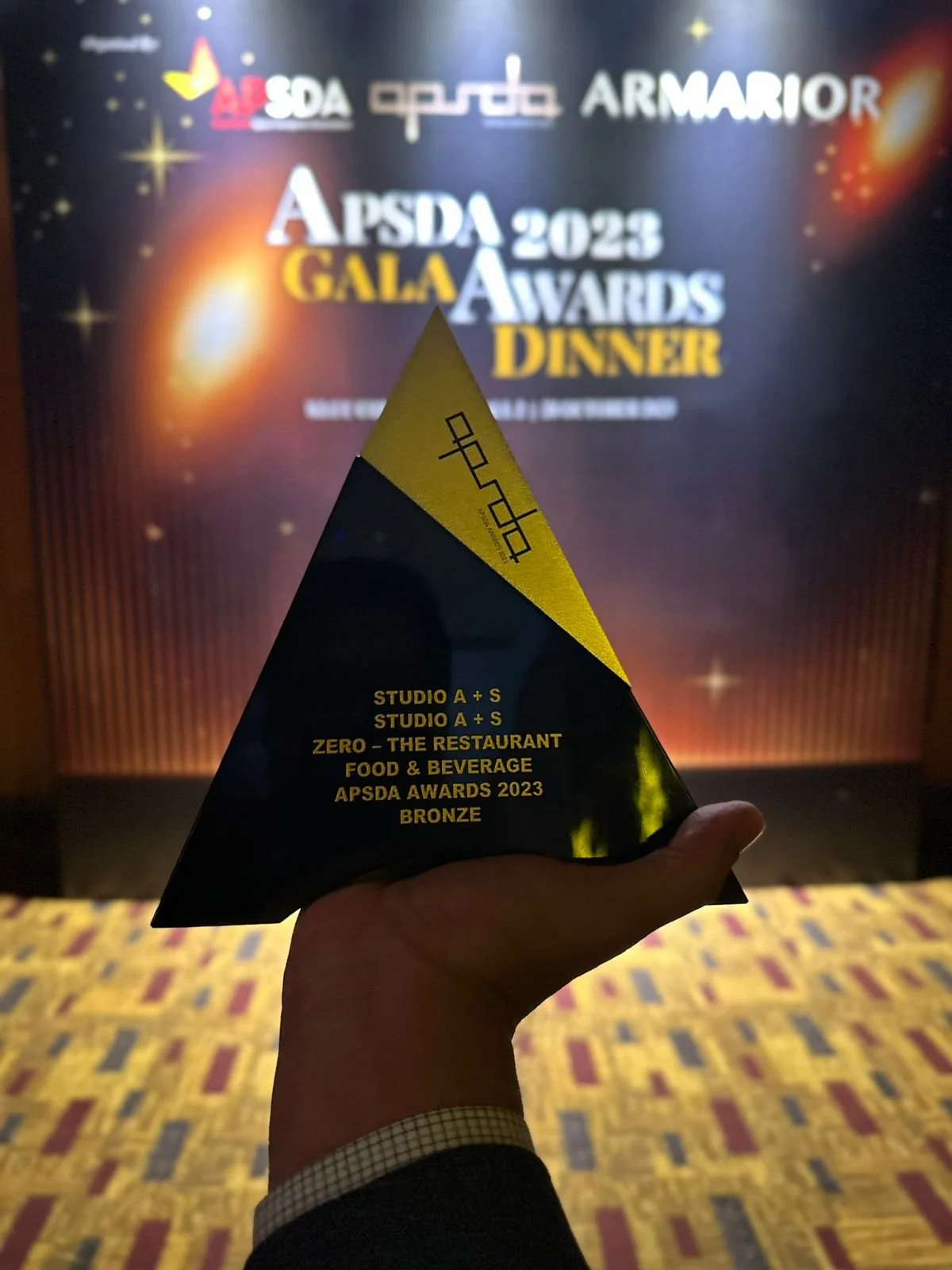 A person holding a black and gold angled award trophy with text on it. The background shows a stage backdrop with the APSDA 2023 Gala Awards Dinner logo and event details.