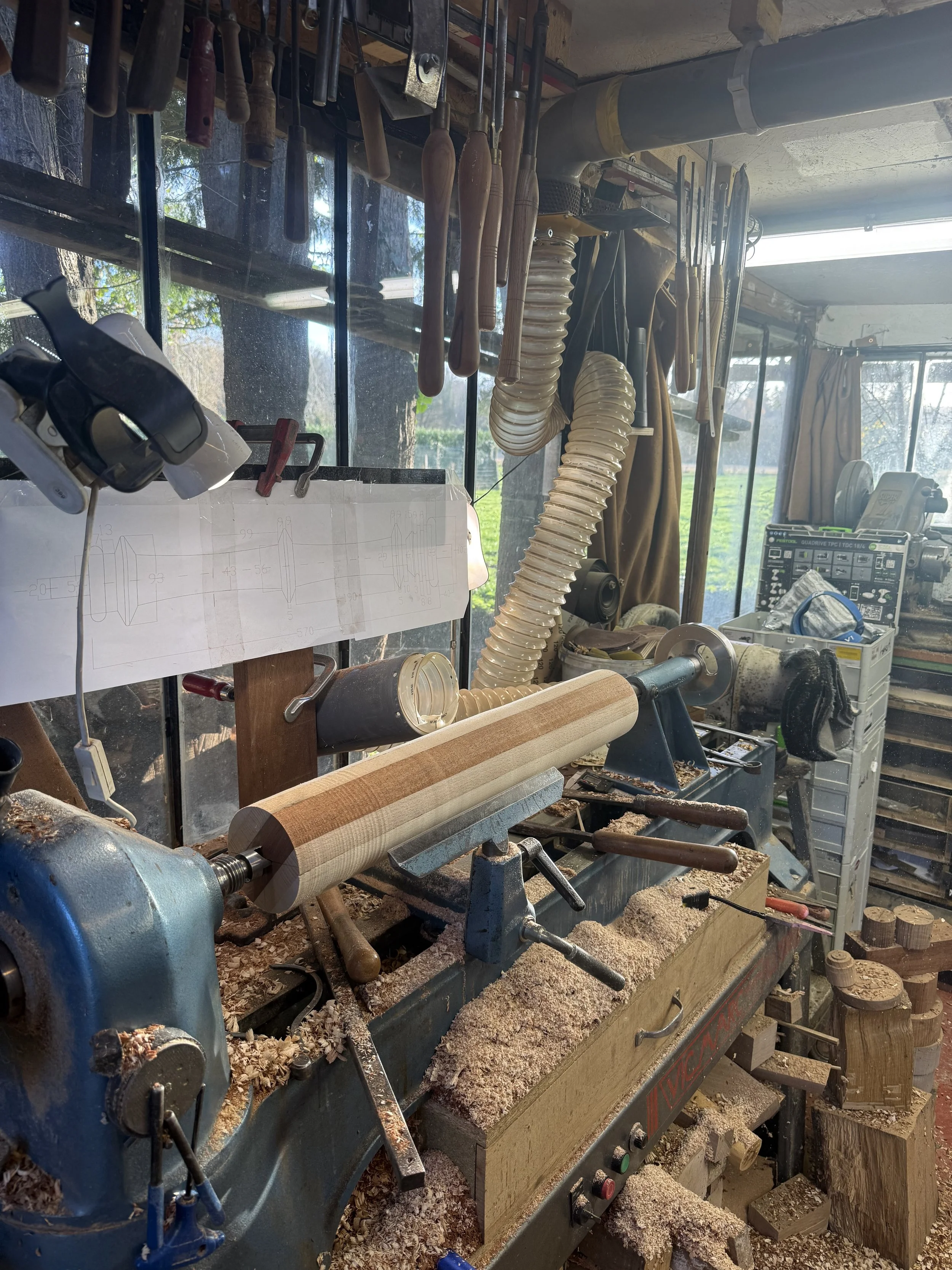 Atelier de tournage sur bois avec un tour mécanique en gros plan, outils suspendus au mur, plan de pièce imprimé, et surface couverte de copeaux de bois.