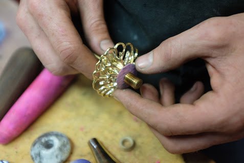 Une personne ajustant une pièce de bijouterie dorée avec une pince, sur un établi de travail.