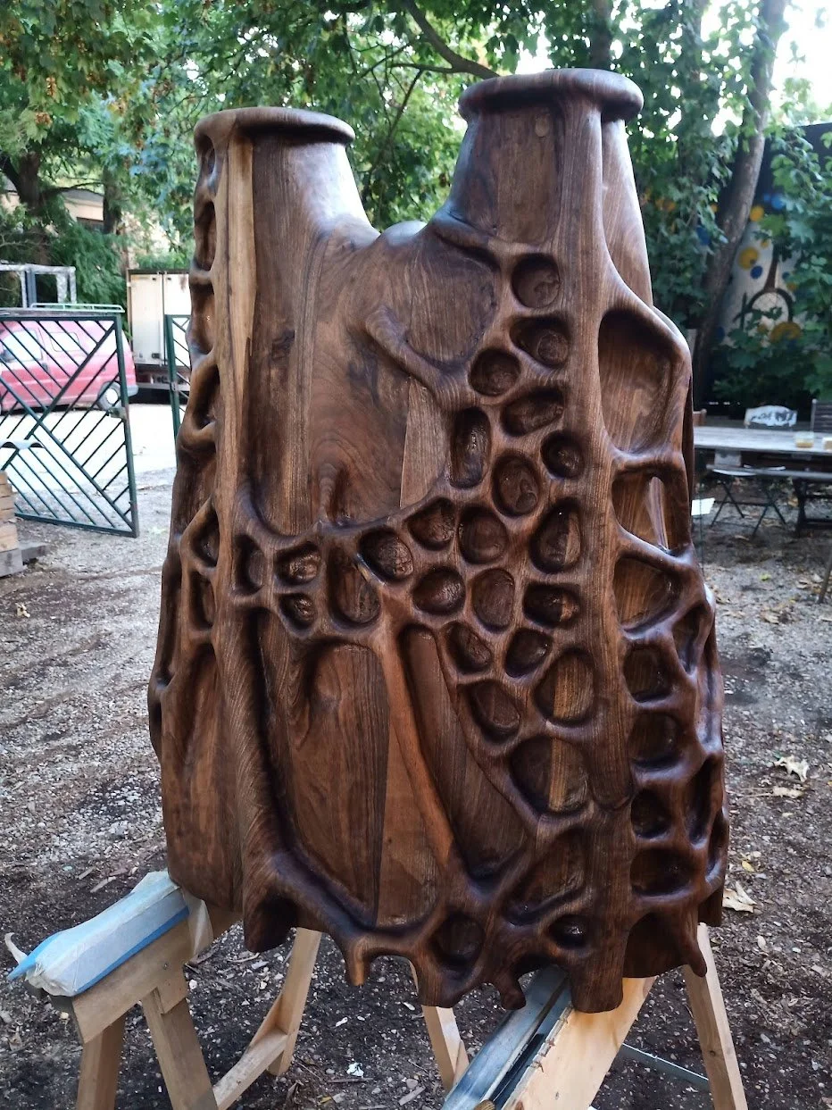 Une sculpture en bois avec un design organique et creusé, située à l'extérieur, entourée d'arbres et d'autres installations.