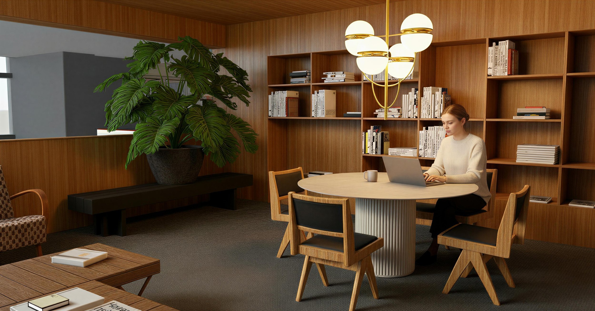 A woman working on a laptop at a round wooden table in a modern office or library, with a large potted plant nearby and wooden shelving filled with books in the background.