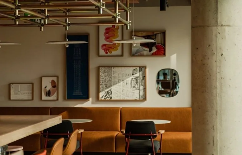 Interior of a modern cafe with artwork on the wall, a tan cushioned bench, black chairs, white round tables, and hanging pendant lights.