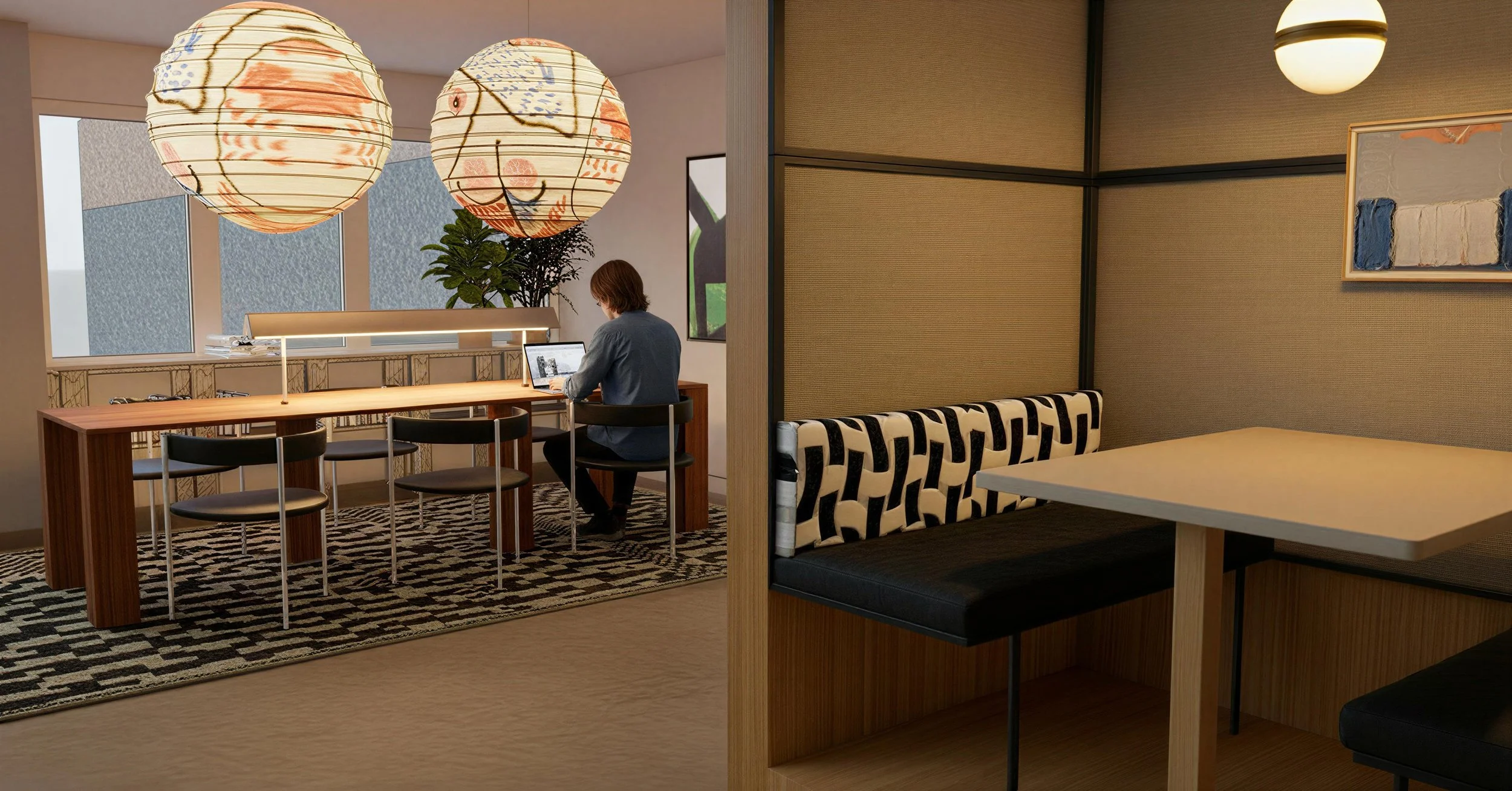 Interior of a modern restaurant or cafe with a booth on the right and a communal table on the left. A person is working on a laptop at the table, illuminated by large decorative paper lanterns hanging from the ceiling. The decor includes patterned cushions, a black-and-white rug, and framed artwork on the walls.