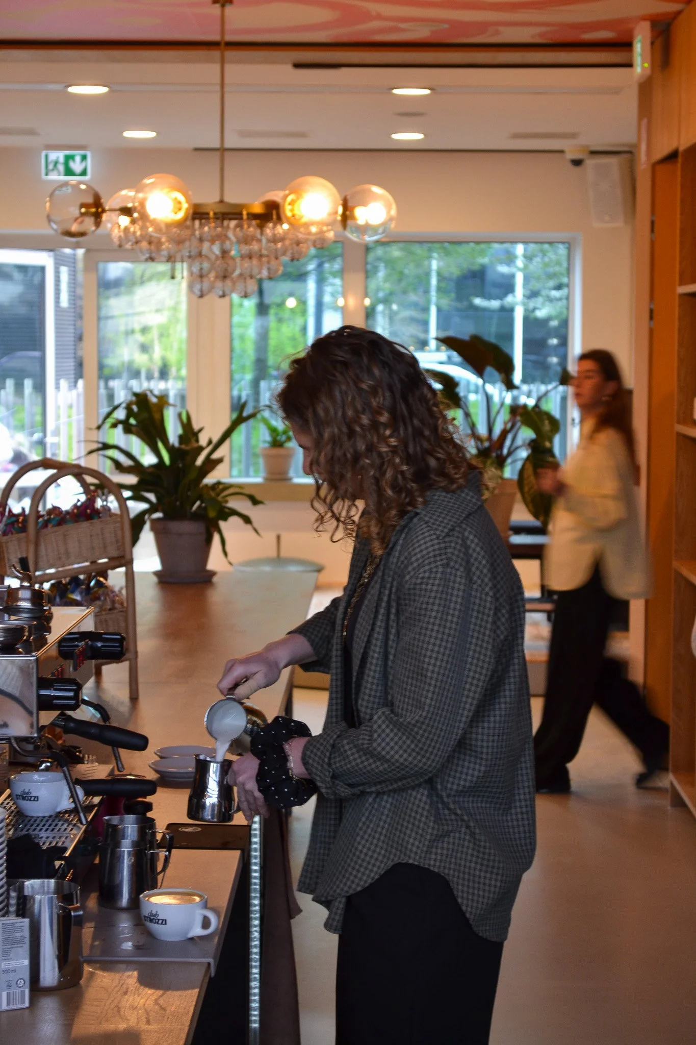 A woman with curly hair wearing a checkered blazer pours a beverage from a container into a white mug at a coffee station inside a well-lit cafe, with another woman in the background by a large window.