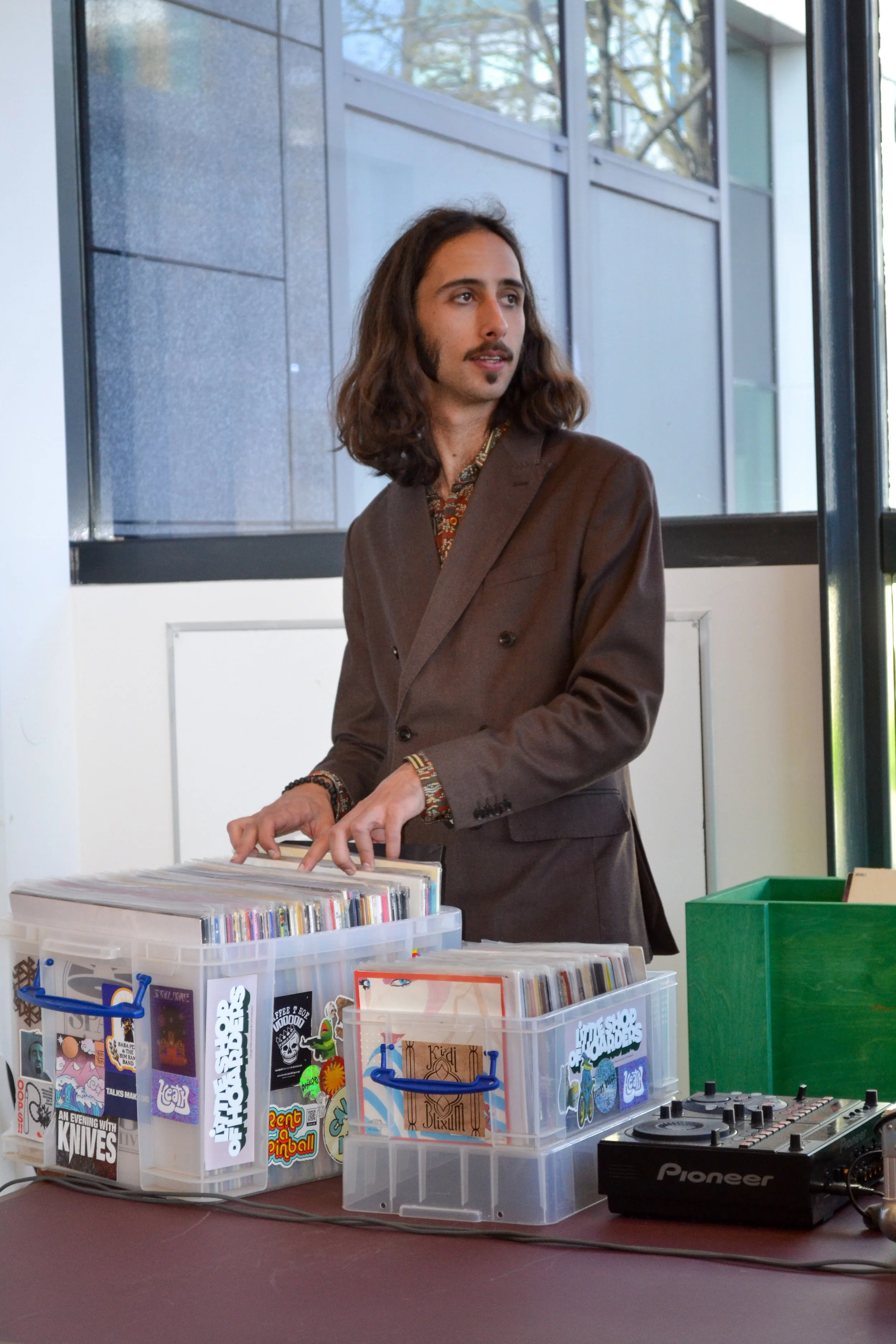 A man with shoulder-length brown hair and a mustache is wearing a brown blazer and colorful shirt. He appears to be playing a keyboard or piano at a table with vinyl records and DJ equipment in a modern indoor space with large windows and natural light.