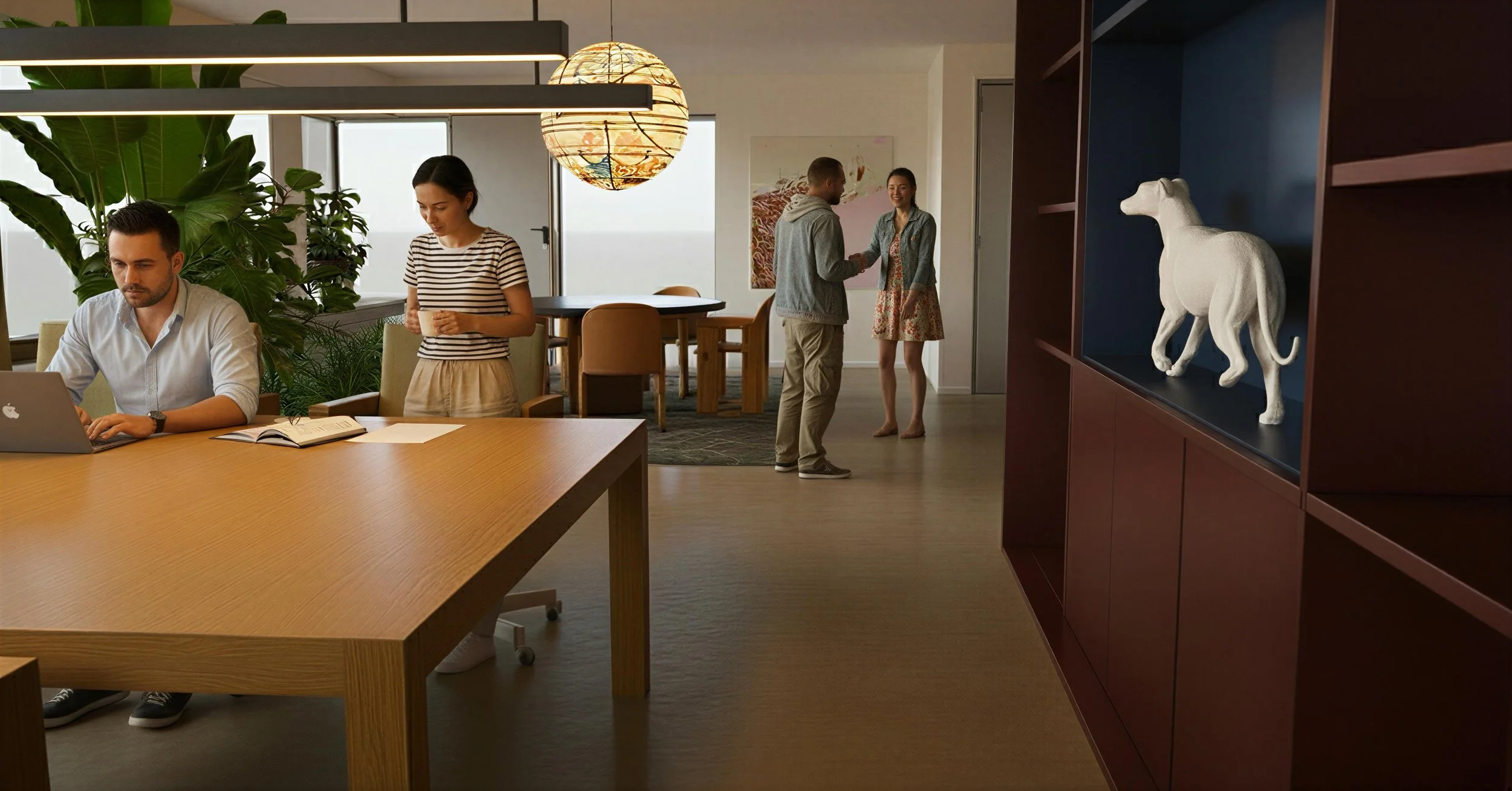 Four people in an office or common area; two men and two women. One man is working on a laptop at a large wooden table, a woman is standing with a cup, and the two other people are standing and chatting near the back.