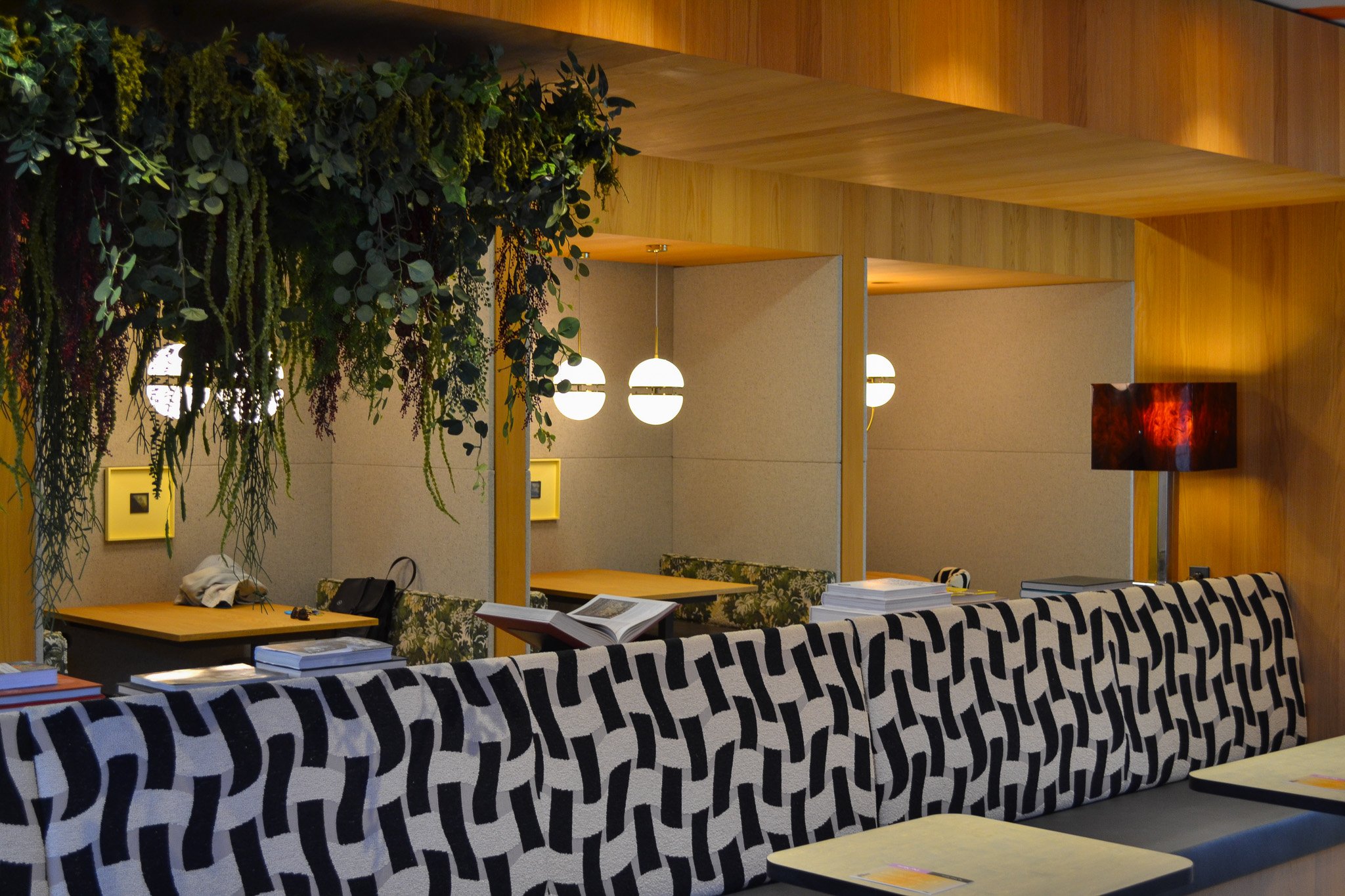 Interior view of a modern lounge or waiting area with wooden walls, hanging lights, a decorated wall with greenery, sofas with patterned covers, tables with books, and a TV screen on the floor