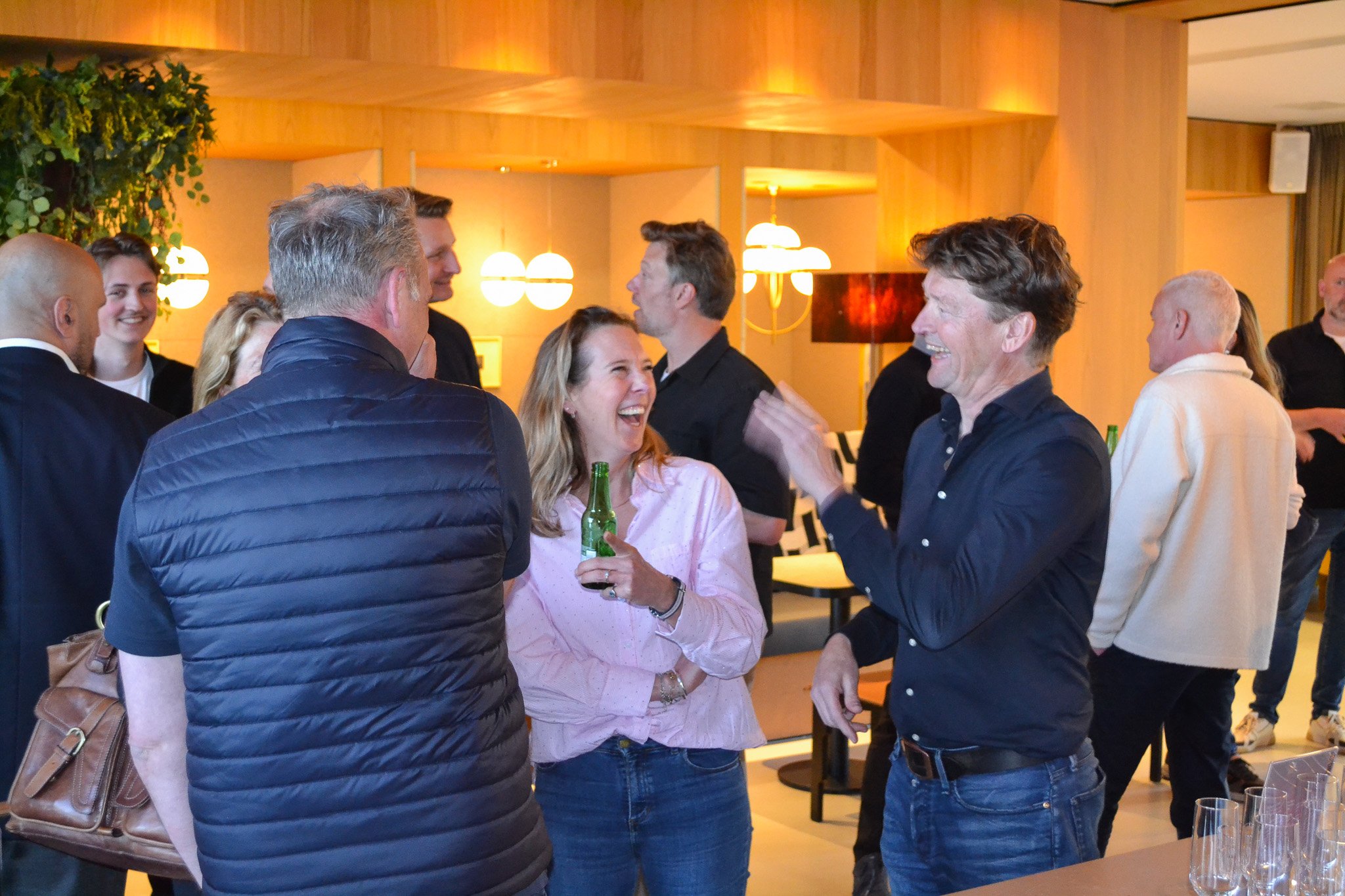 People gathered indoors, laughing and talking, with some holding drinks, in a warmly lit room with wooden walls and modern lighting fixtures.