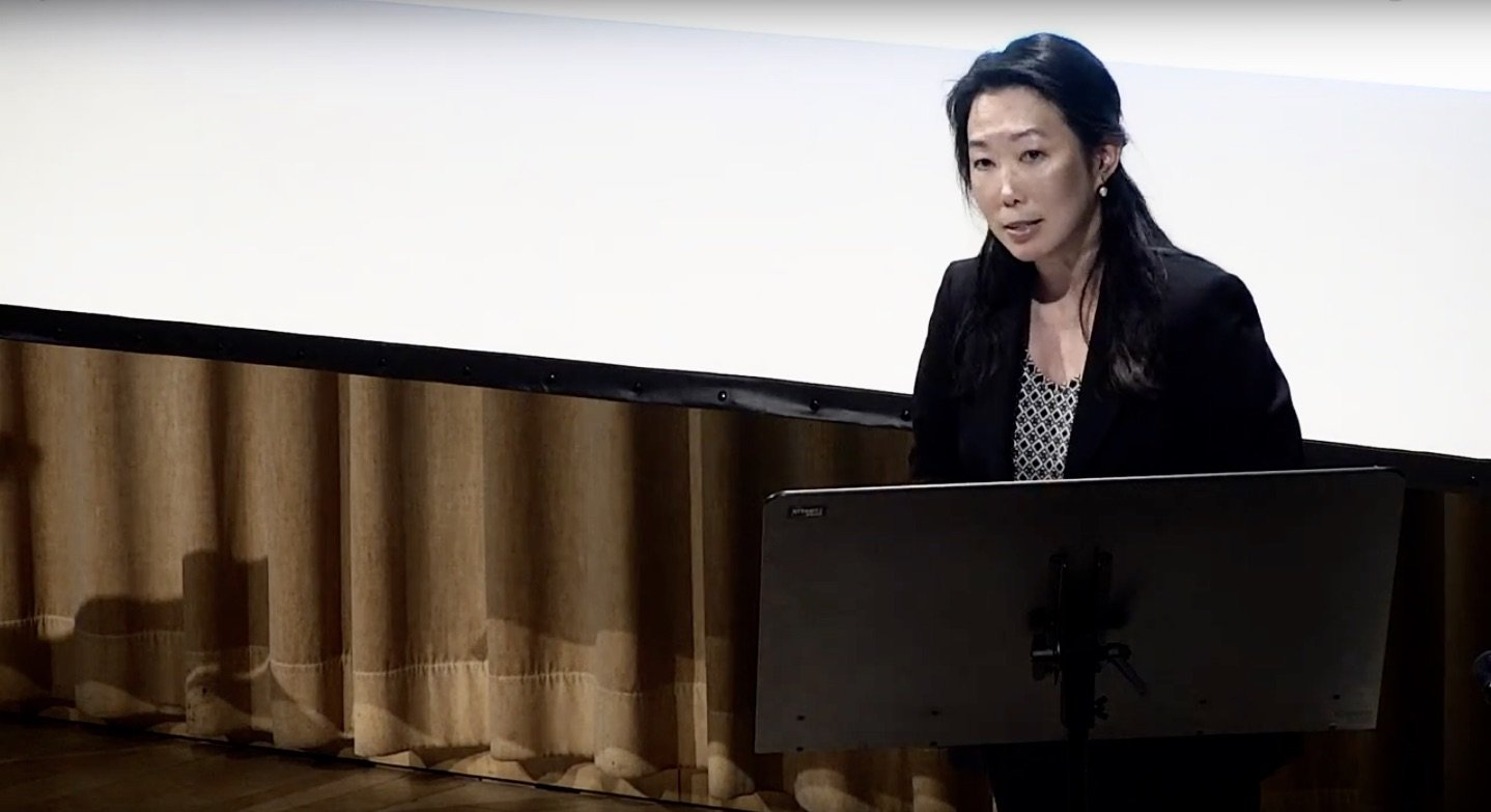 A professional woman lecturing in a black blazer over a patterned blouse, is sitting behind a laptop in front of a white projection screen and brown curtains.