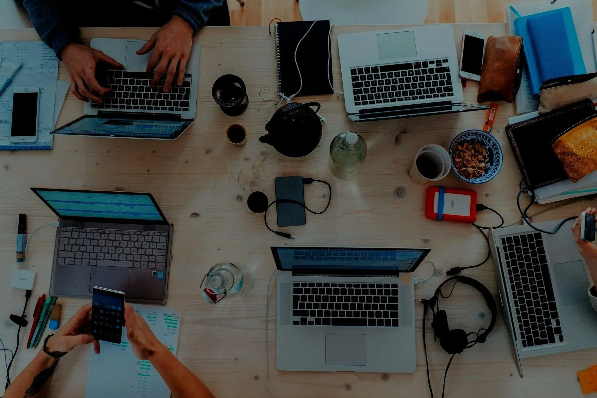 A top-down view of a cluttered wooden table with multiple laptops, smartphones, notebooks, drinks, and various office supplies, with hands of people working on different devices.