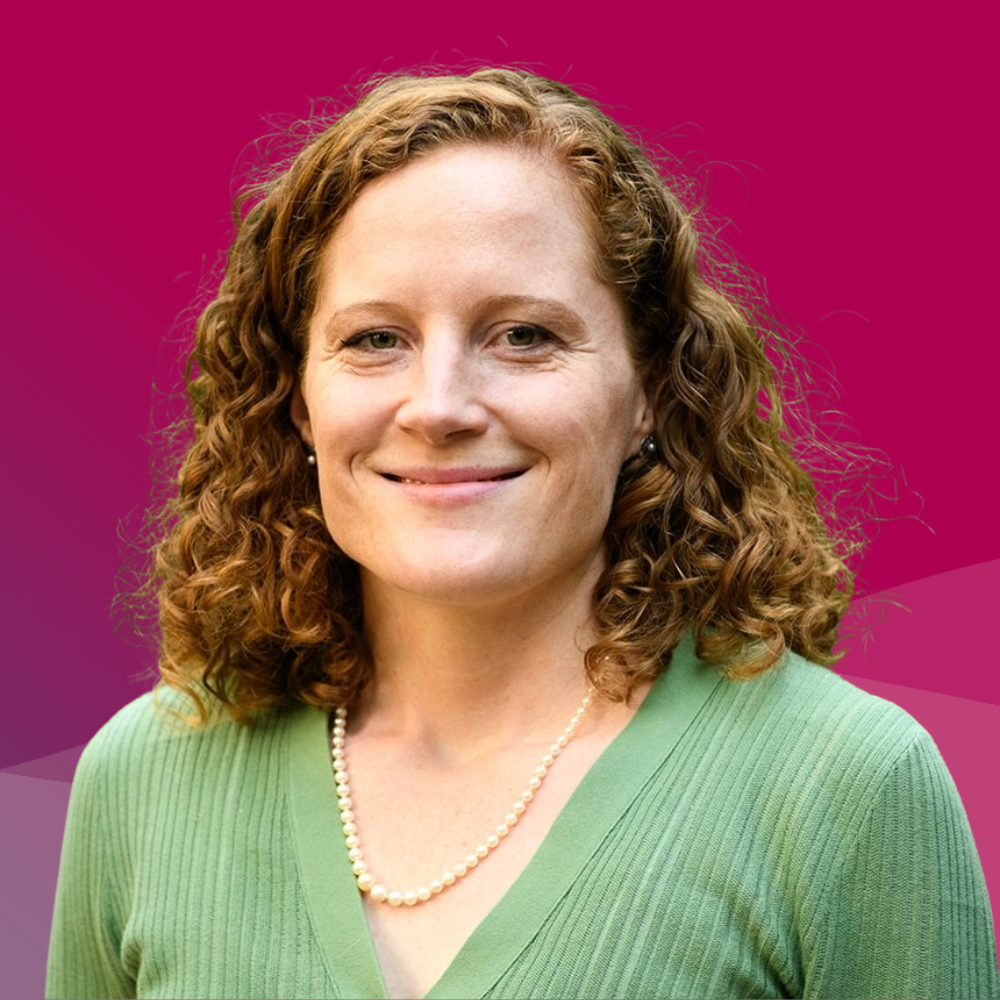 A woman with shoulder length red hair and a green cardigan smiles at the camera with a raspberry colored background.