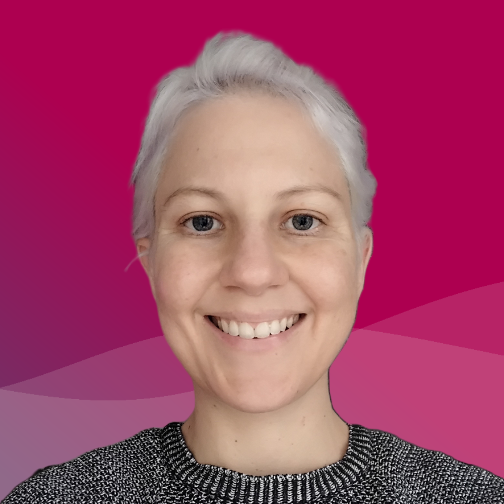 A woman with silver colored hair and a black and white sweater looks straight at the camera with a bright smile and against a raspberry background.