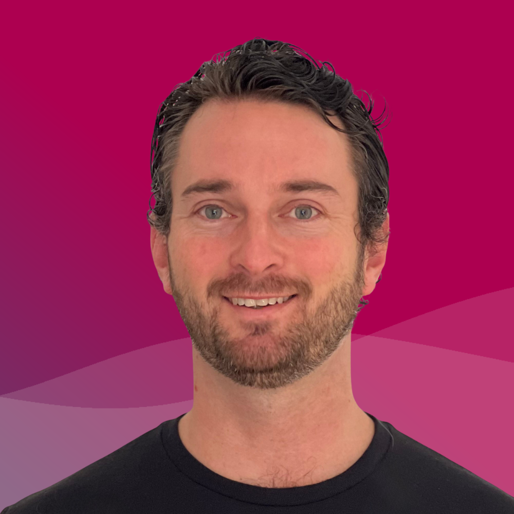 A man with dark hair, a beard, and blue eyes is smiling at the camera with a pink and purple abstract background.