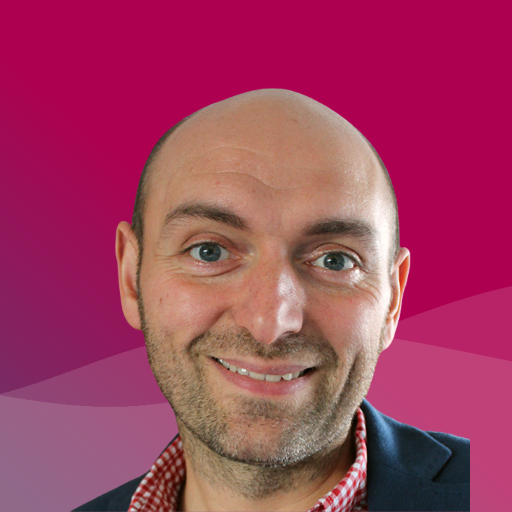 A close-up of a smiling man with a bald head, wearing a dark blazer and a red checkered shirt, against a pink and purple gradient background.