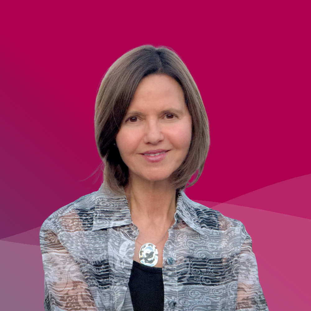 A woman with shoulder-length brown hair smiling, wearing a patterned button-up shirt and a skull pendant necklace, against a pink and purple gradient background.