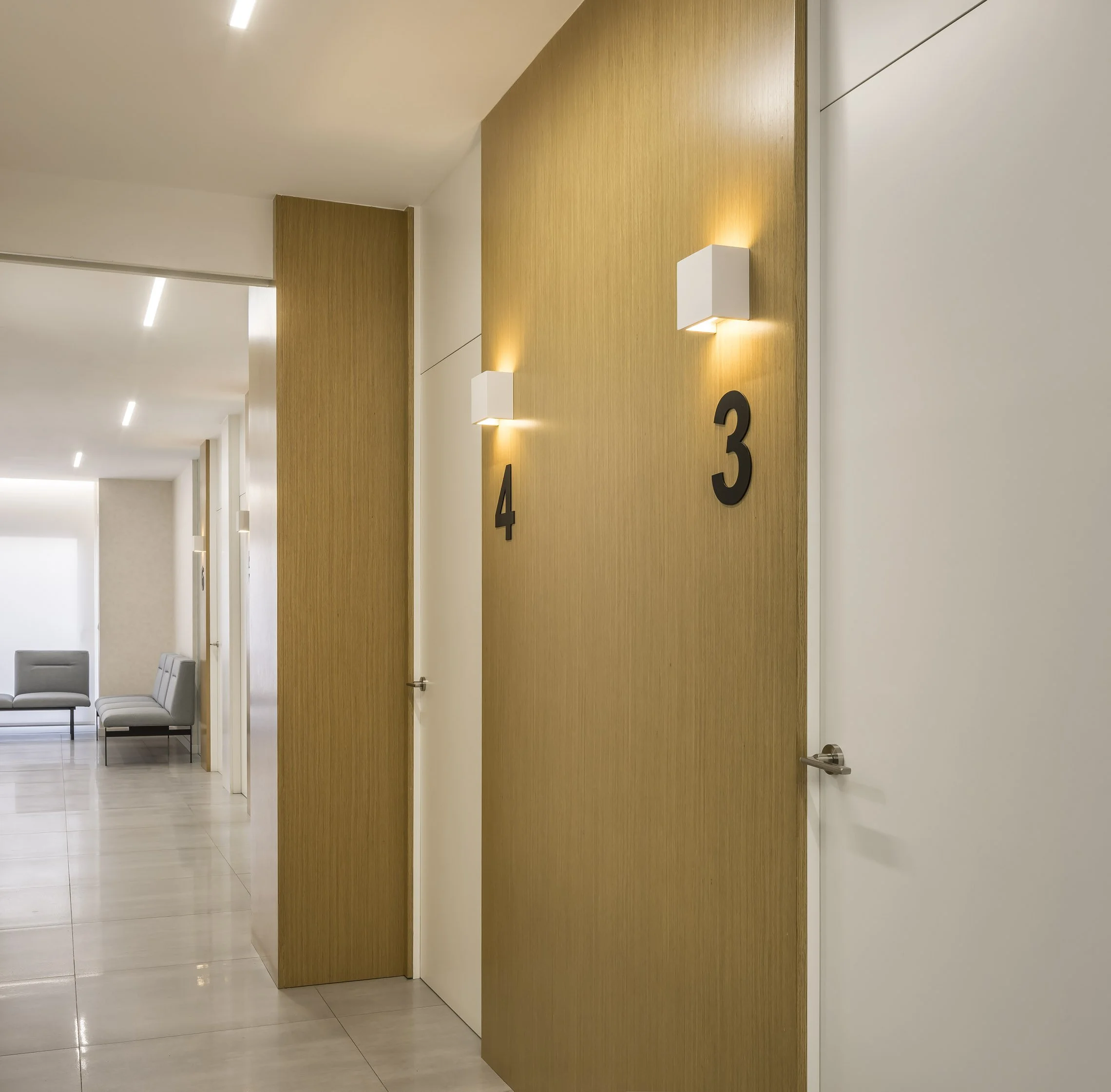 Pasillo de un edificio con paredes blancas y una pared de madera con números 3 y 4, sillones de espera y puertas cerradas.