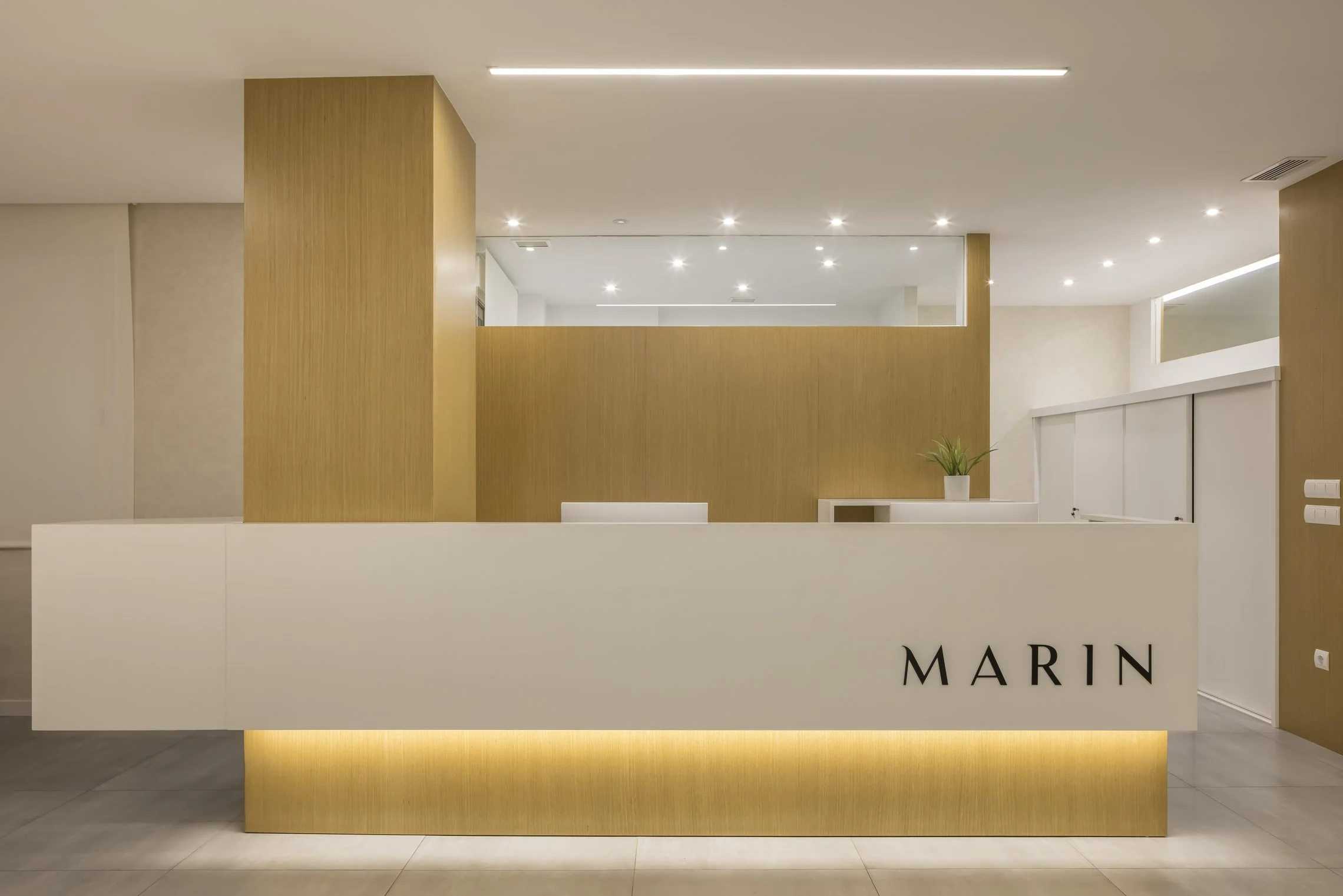 Mostrador de recepción en un ambiente moderno con paredes de color madera y luminarias empotradas en el techo, con planta decorativa y la palabra 'MARIN' en el frente.