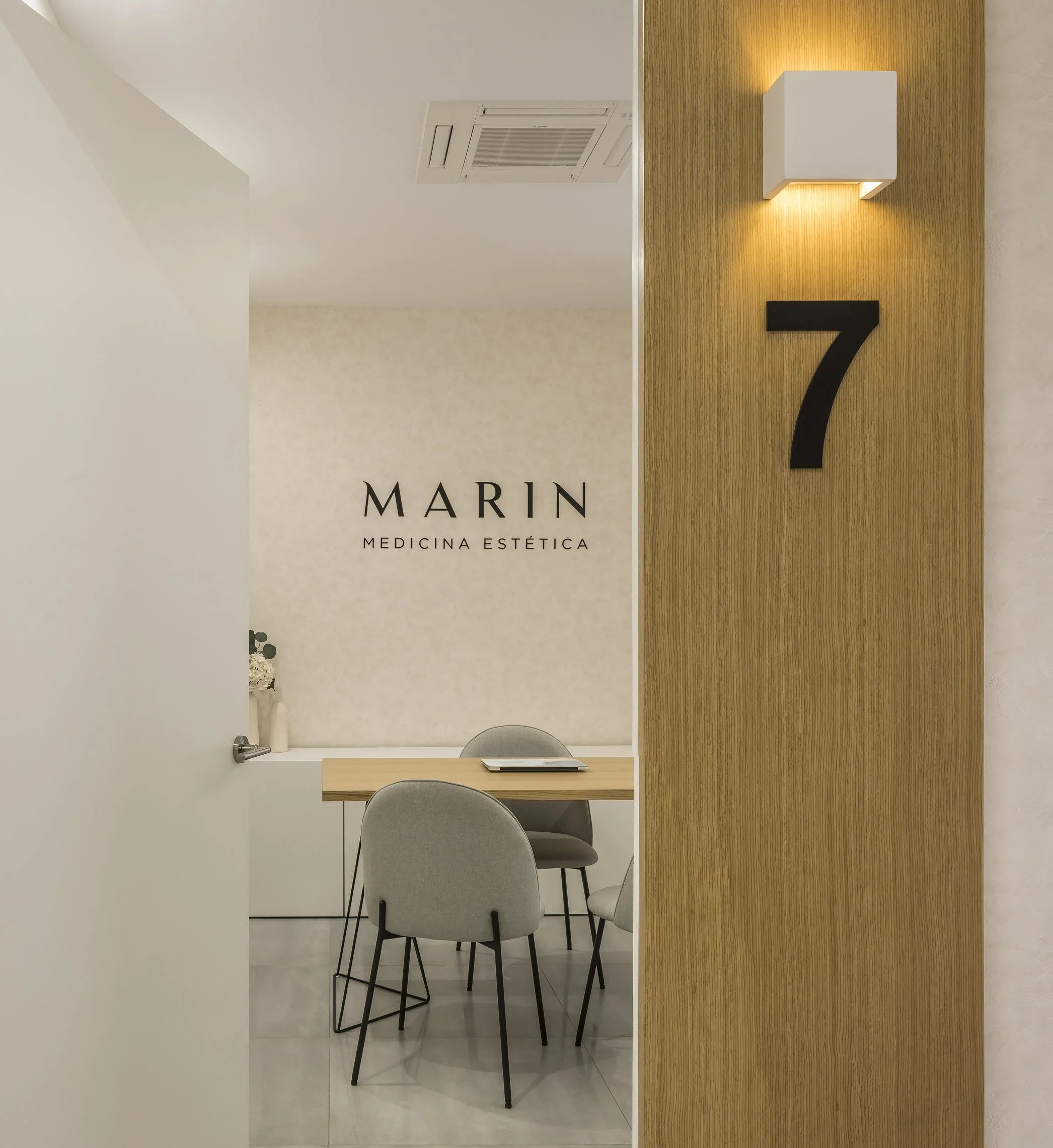 Área interior de una clínica estética con una mesa de madera, cuatro sillas grises, una pared de madera y un cartel que dice 'Marin Medicina Estética'.