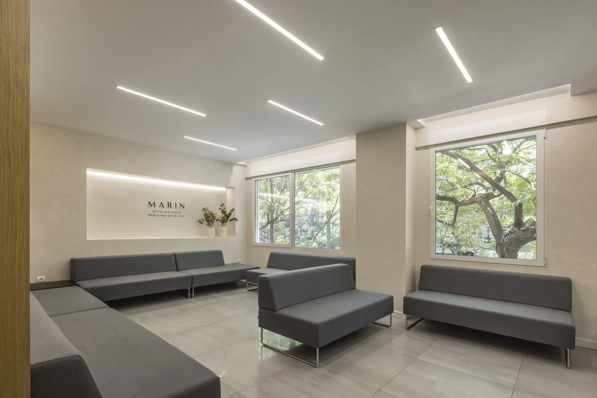 Sala de espera moderna con sillones grises, ventanas grandes con vista a árboles verdes, paredes blancas y techo con luces lineales.