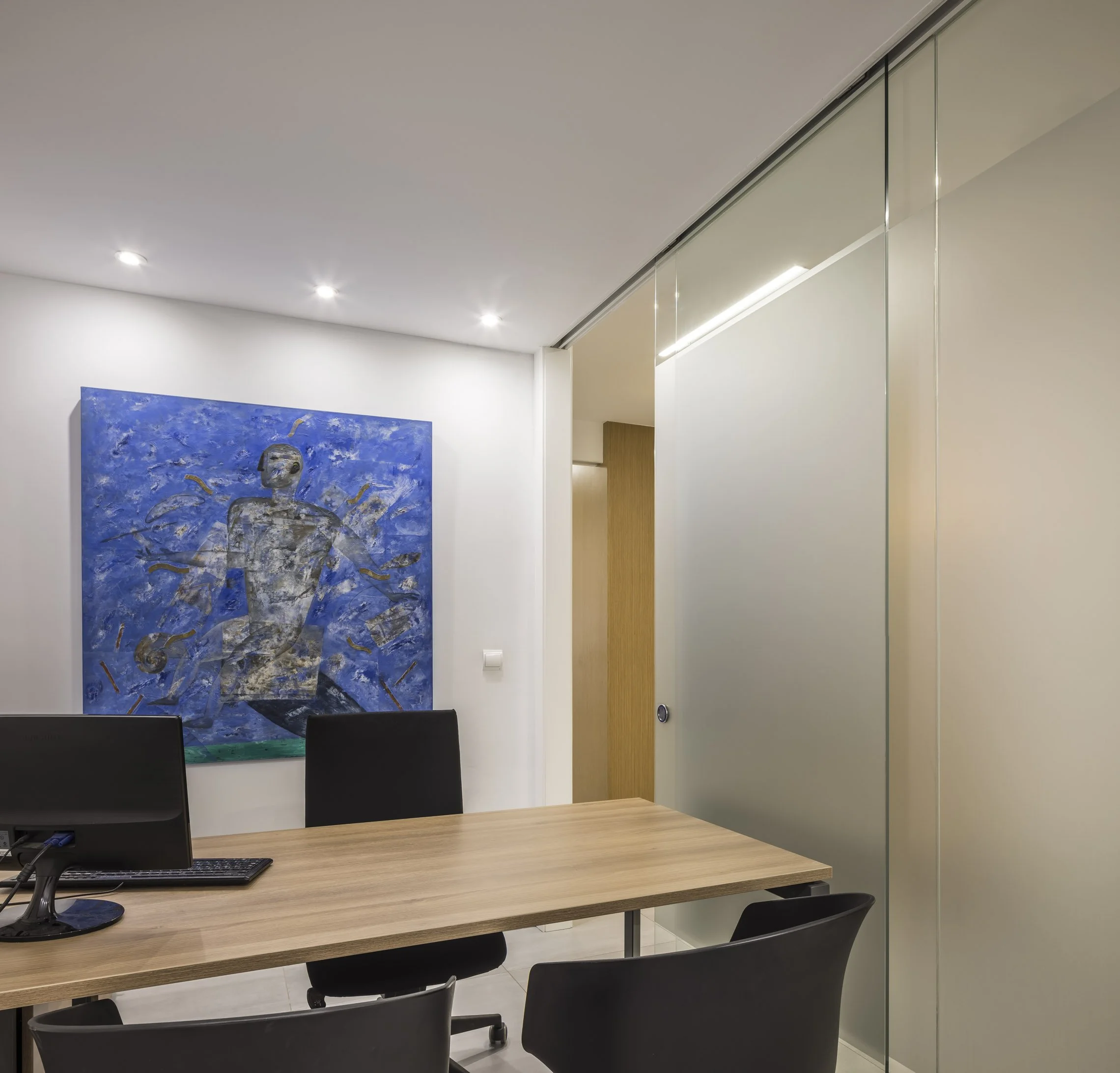 Sala de reuniones moderna con mesa de madera y sillas negras, paredes blancas, cuadro azul y puerta de vidrio con marco de aluminio.