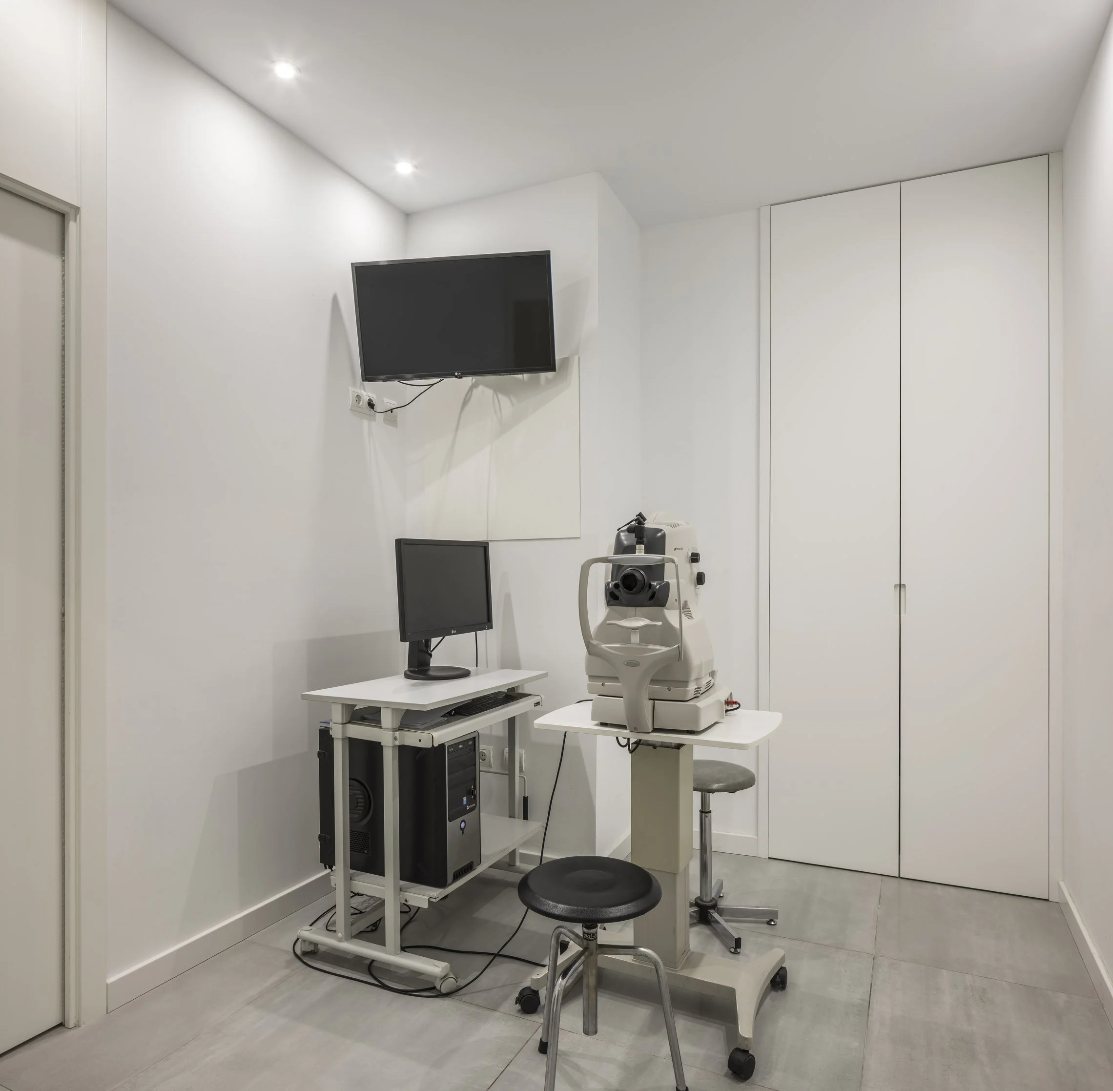 Sala de consulta médica con equipo de tecnología, incluyendo monitor, computadora y equipo oftalmológico, en una habitación blanca y moderna.