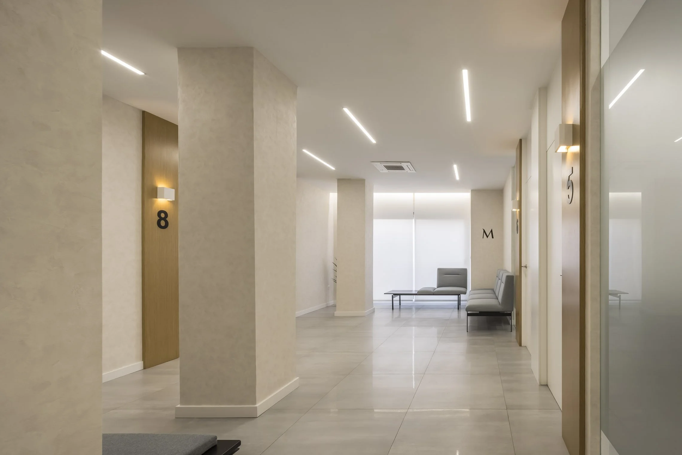 Pasillo de edificio con paredes beige, puertas con números, sillones y mesas de espera, iluminación moderna en el techo.