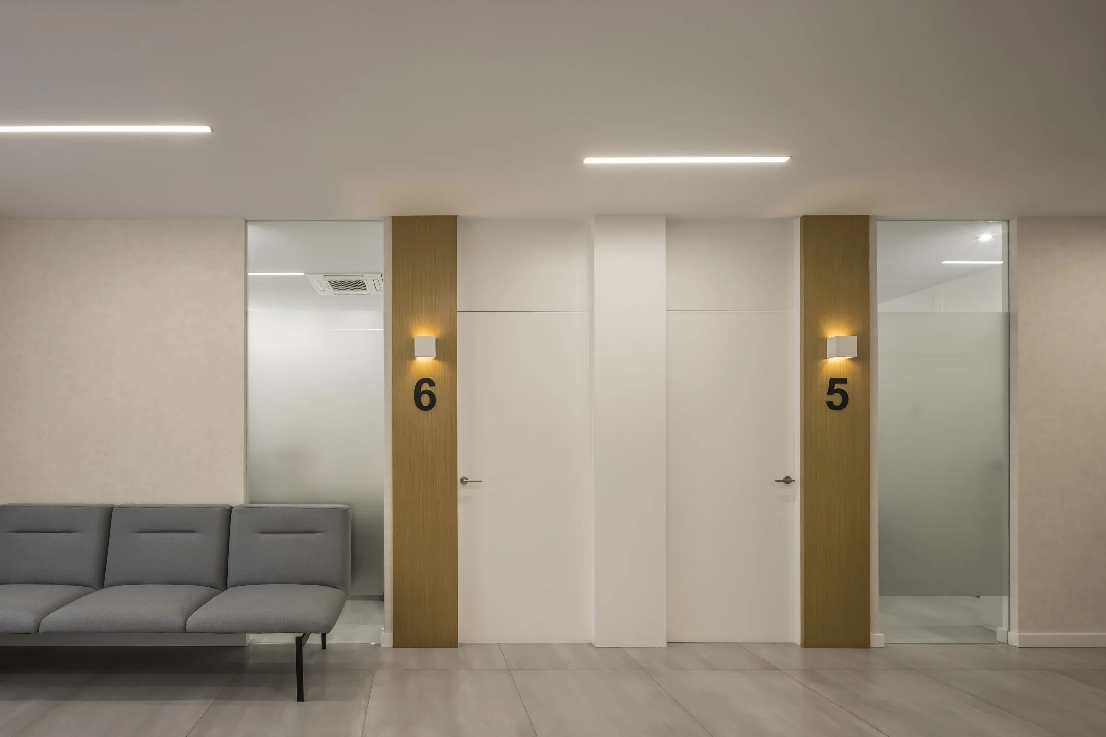 Sala de espera moderna con paredes blancas, dos muros de madera con luces de pared, puertas numeradas 5 y 6, y un sillón gris.