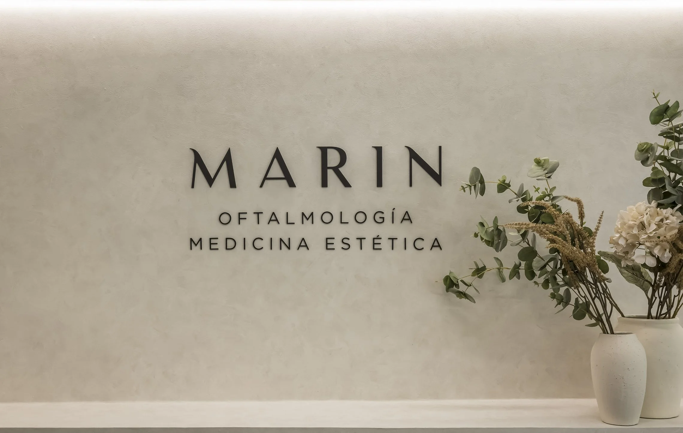 Cartel en la pared que dice 'MARIN', seguido de servicios de oftalmología, medicina estética y otro en letras pequeñas. A la derecha, un jarrón con flores y ramas verdes.